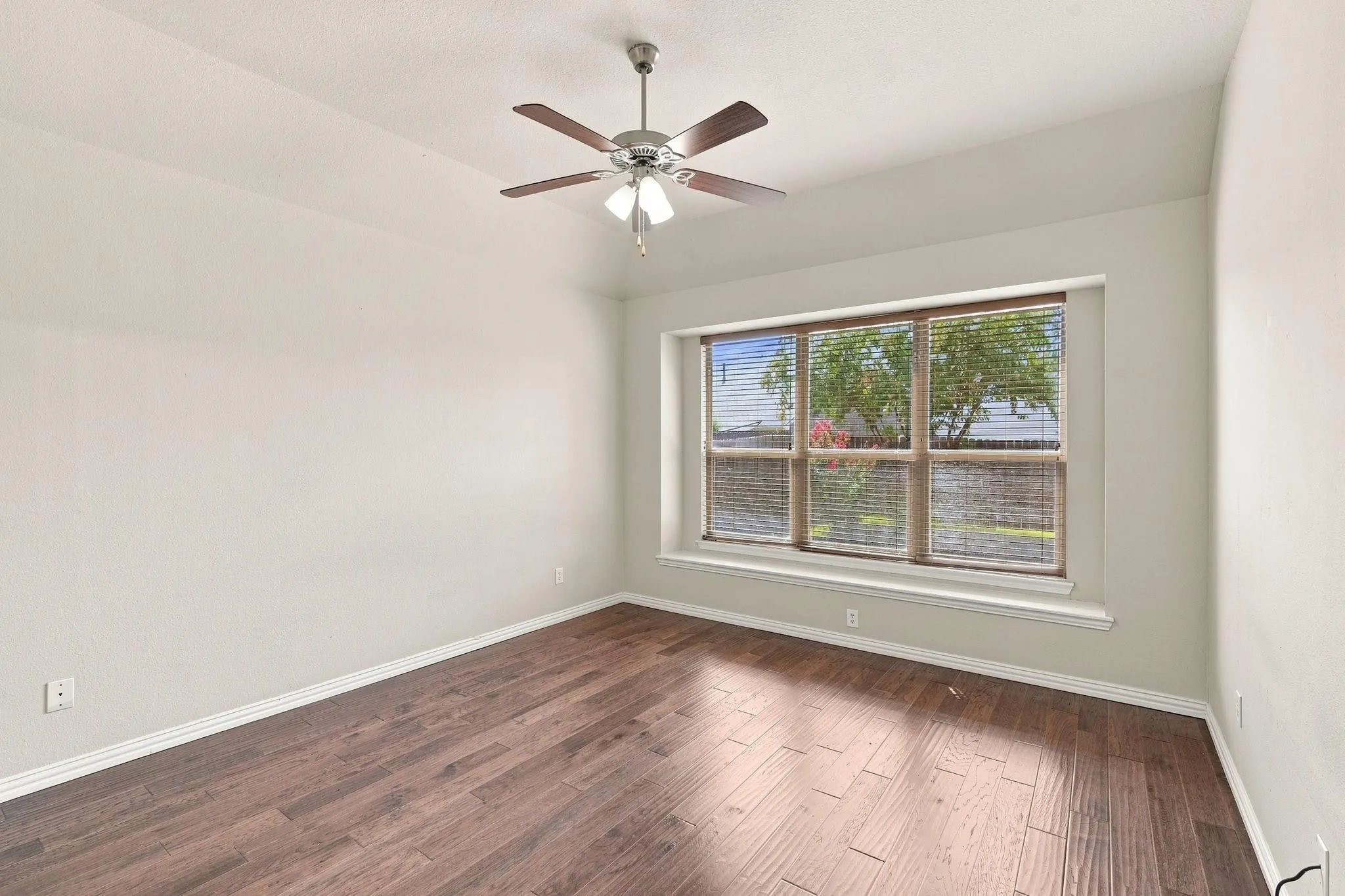 Wood-finish flooring with a neutral wall tone, featuring a ceiling fan with integrated lighting and a large window with blinds
