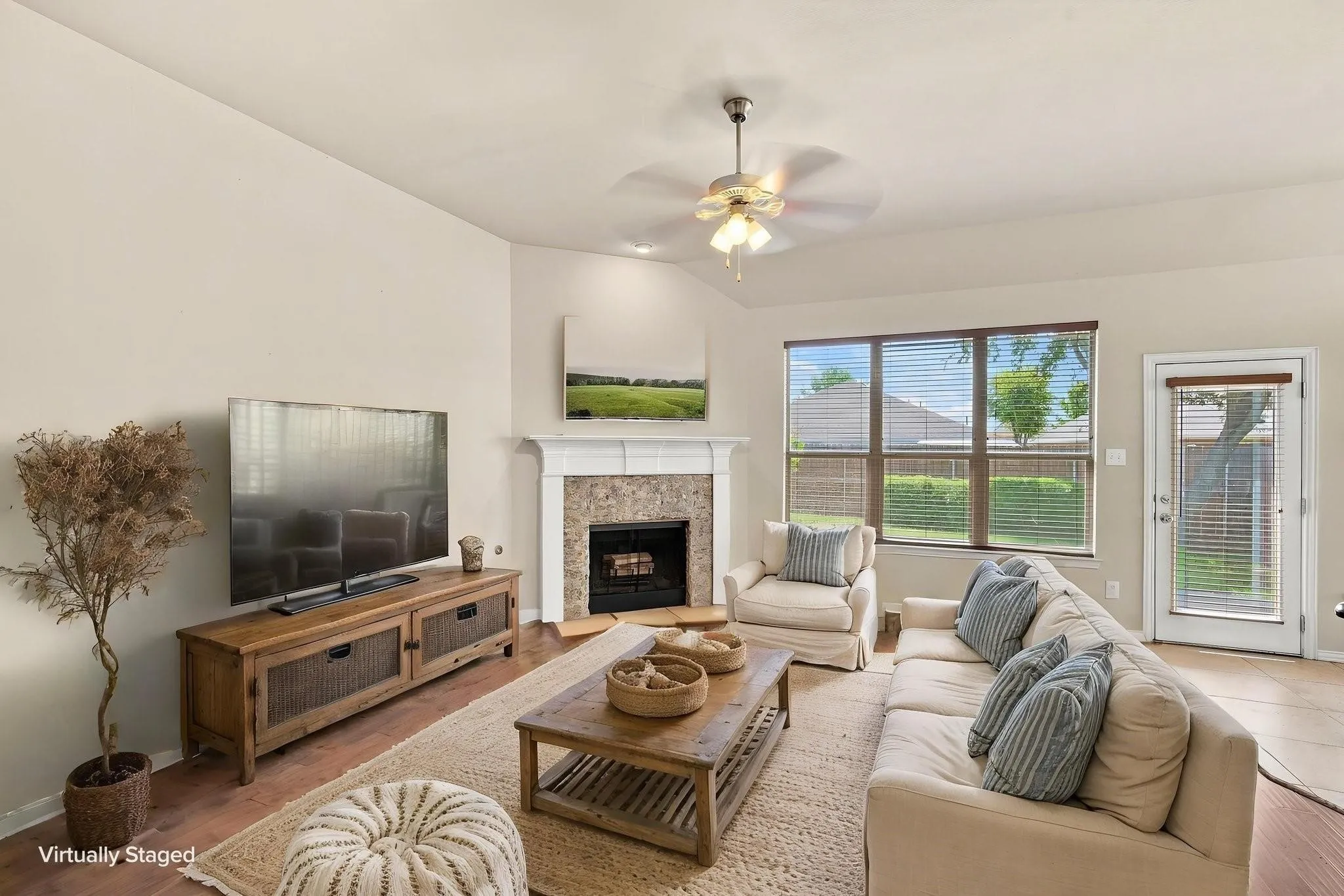Wood-finish flooring, a ceiling fan, a fireplace with a stone surround and white mantel, a large window, and a glass-paneled door