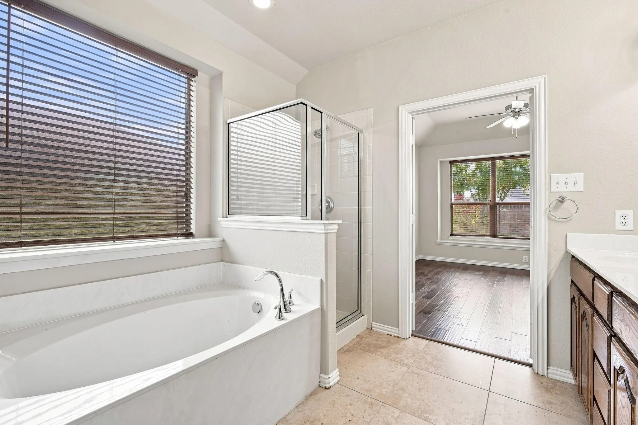 Soaking tub, walk-in shower with glass enclosure, large window with blinds, tile flooring, and wood-finish cabinetry
