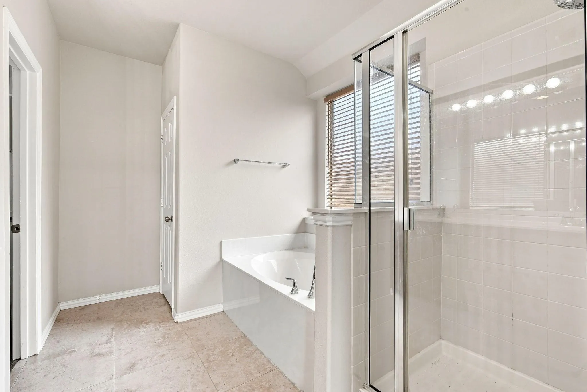 Tiled flooring, built-in bathtub, glass-enclosed shower, white tile shower surround, and window with blinds