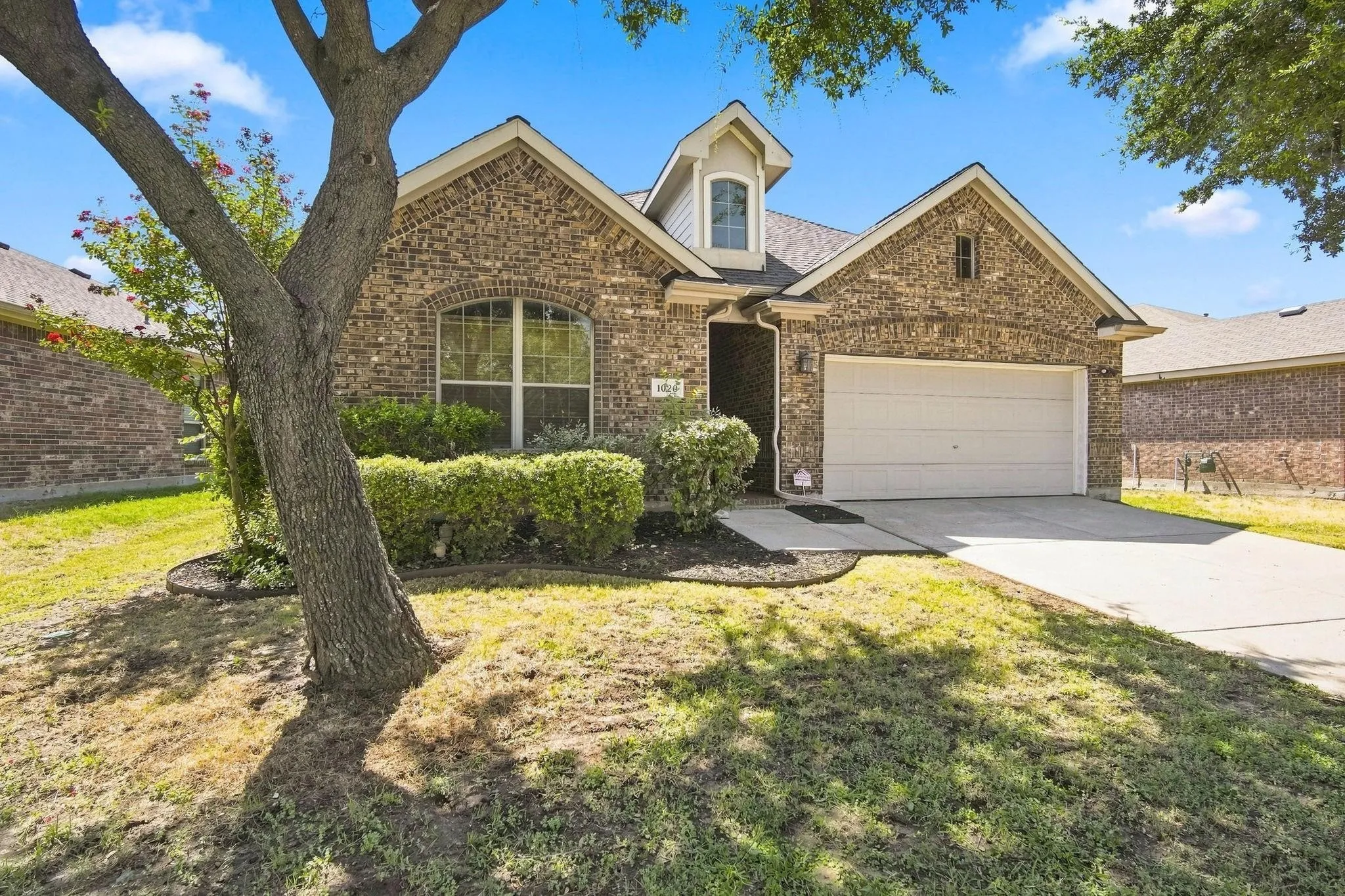Brick exterior with an arched window, dormer architectural element, attached garage, and landscaped front yard