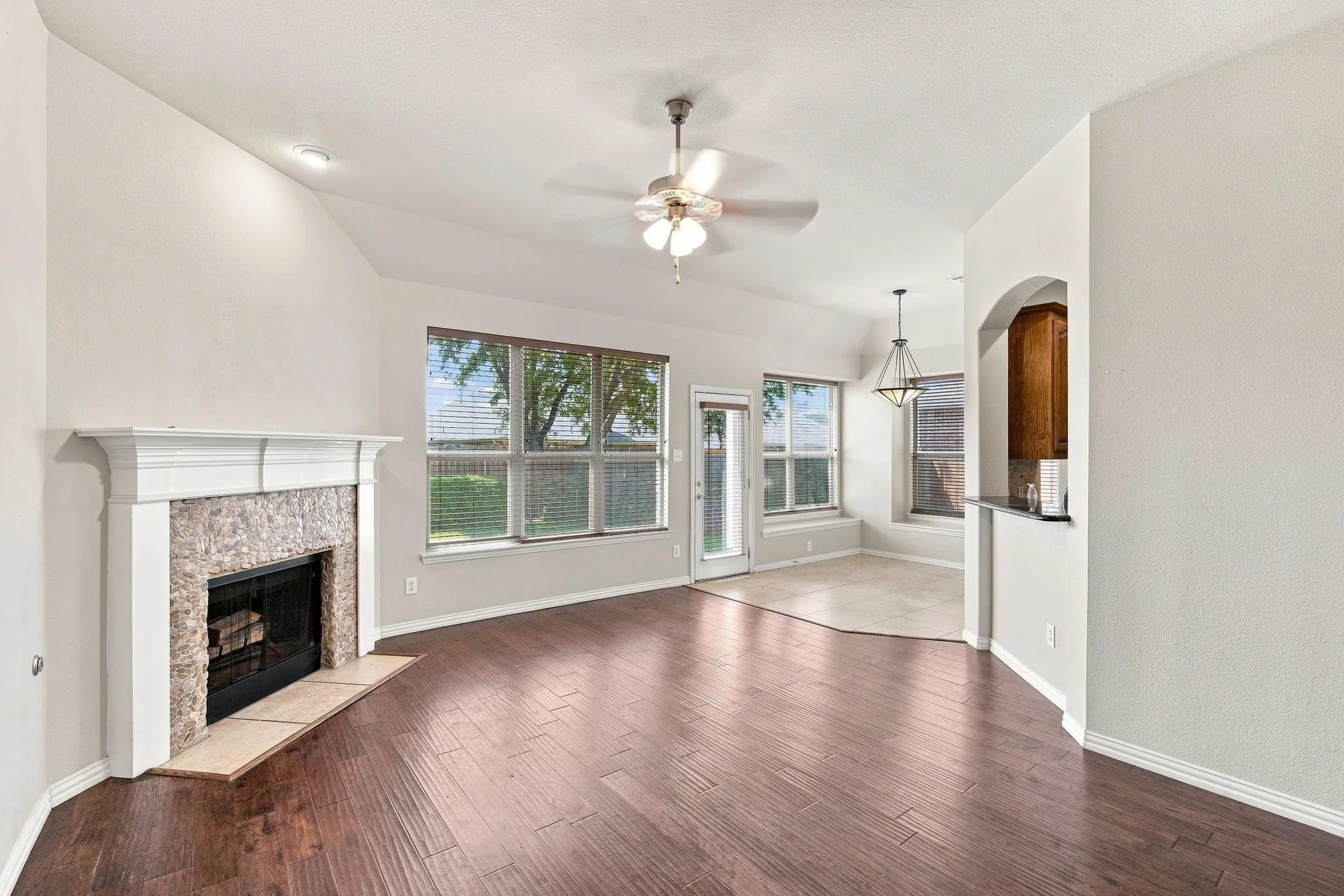 Vaulted ceiling with a ceiling fan, wood-finish flooring, stone-faced fireplace with a mantel, multiple windows, and an arched pass-through to the kitchen