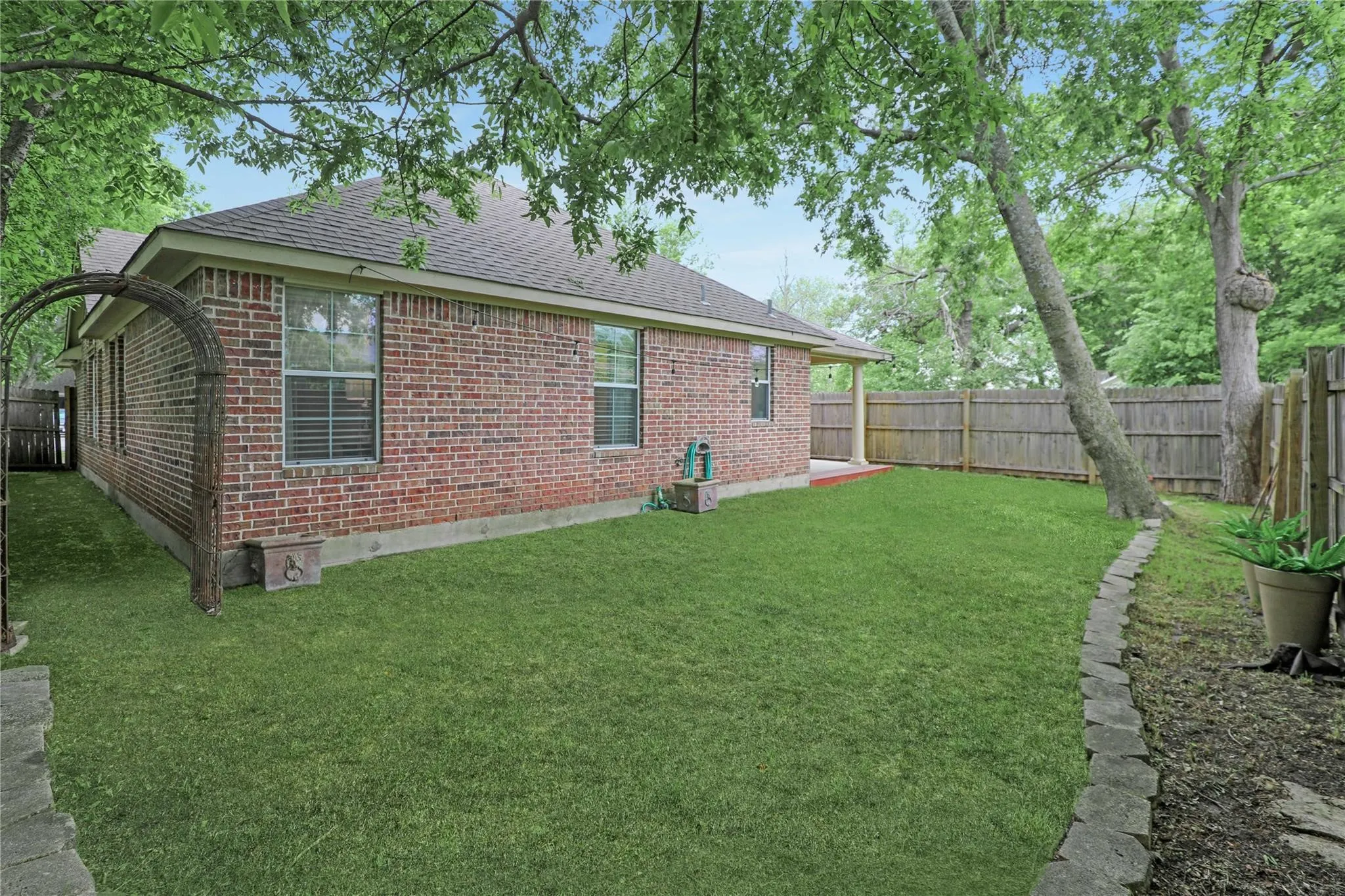 Spacious backyard featuring a brick exterior, shingled roof, mature trees, wood fence, and stone-edged landscaping