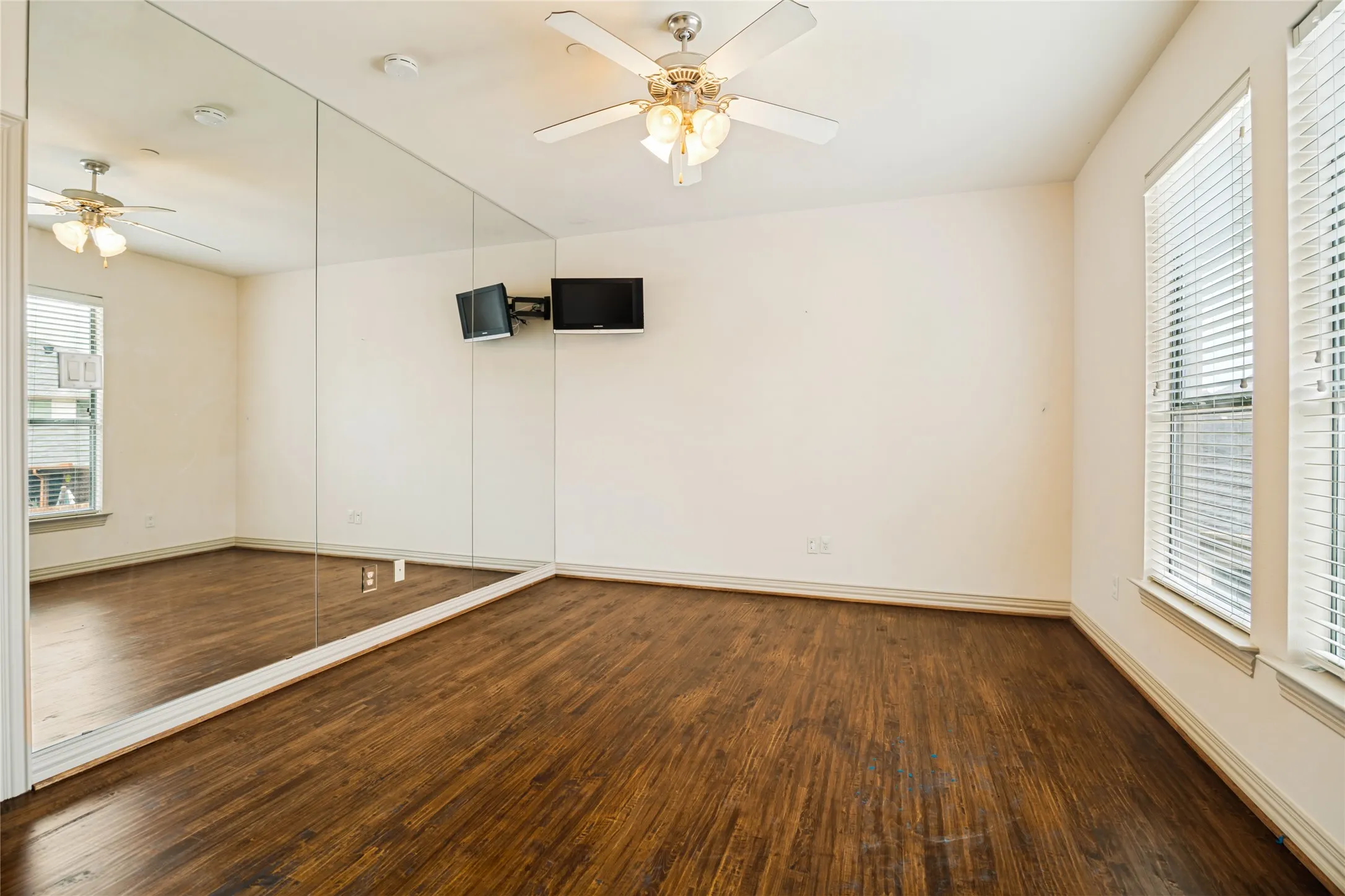 Full-height mirror wall, dark wood-finish flooring, white walls, two windows with blinds, and a ceiling fan with integrated lighting