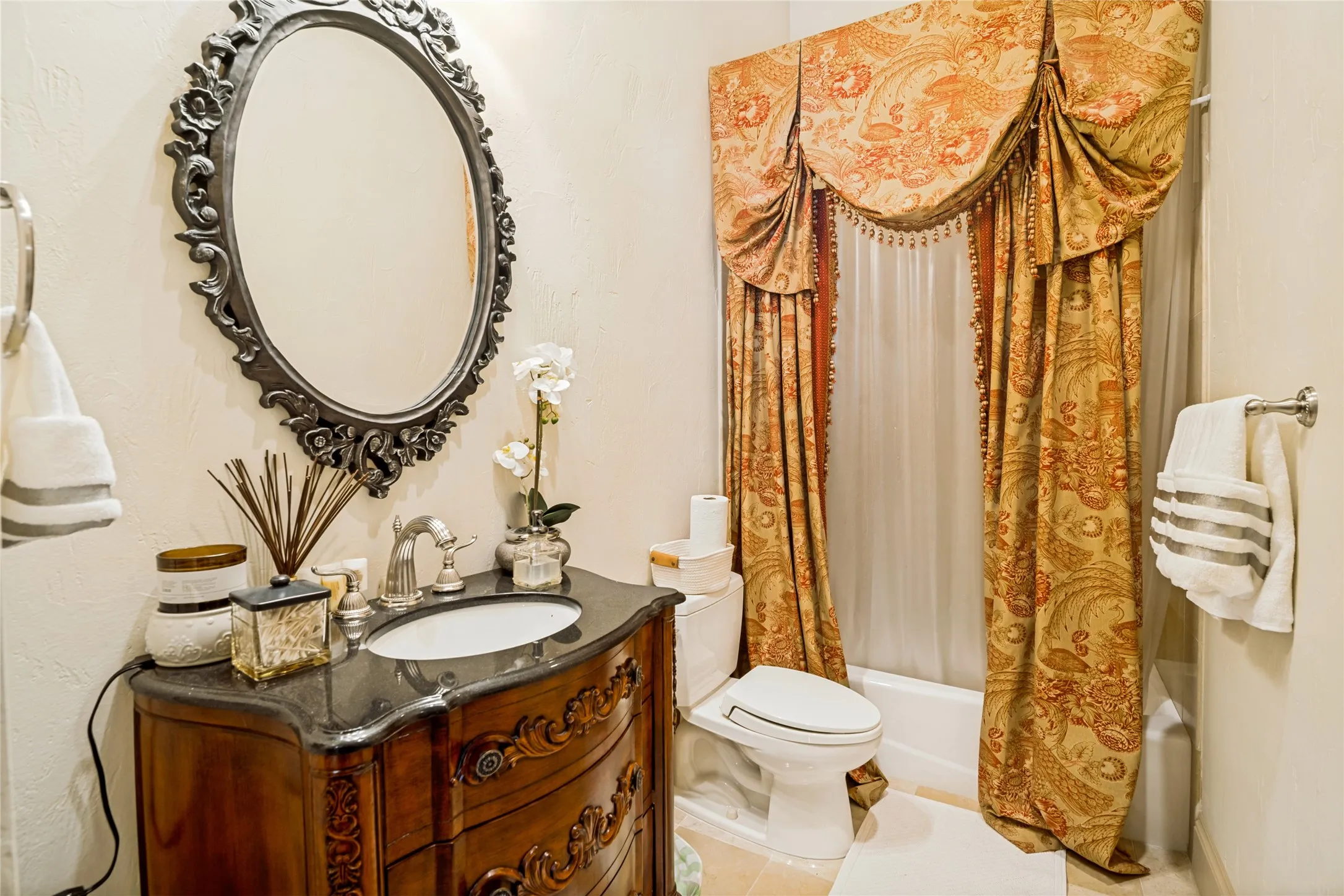 Carved wood vanity with an integral sink, ornate framed oval mirror, and a tub-shower combination with decorative valance