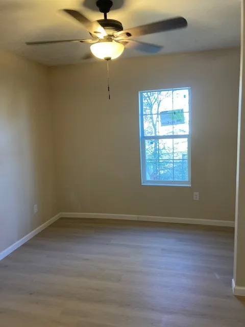 Wood-finish flooring, neutral wall tones, white trim, window, and a ceiling fan with integrated lighting