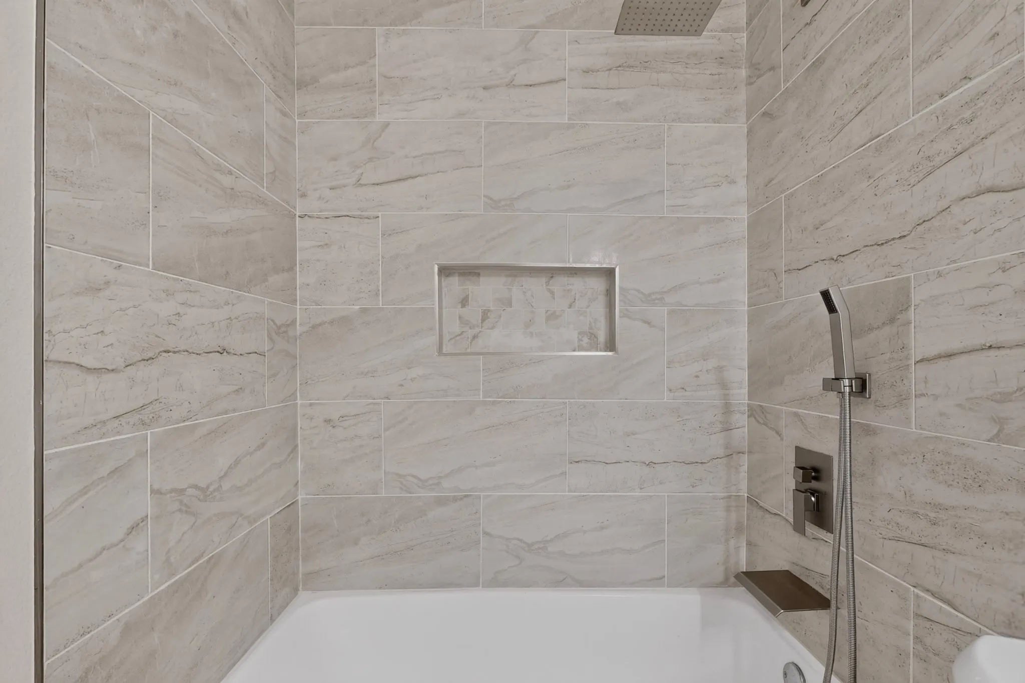 Bathtub with shower system featuring a rainfall showerhead and handheld sprayer, complete with a built-in niche and wood-finish shelf, all surrounded by large format tile
