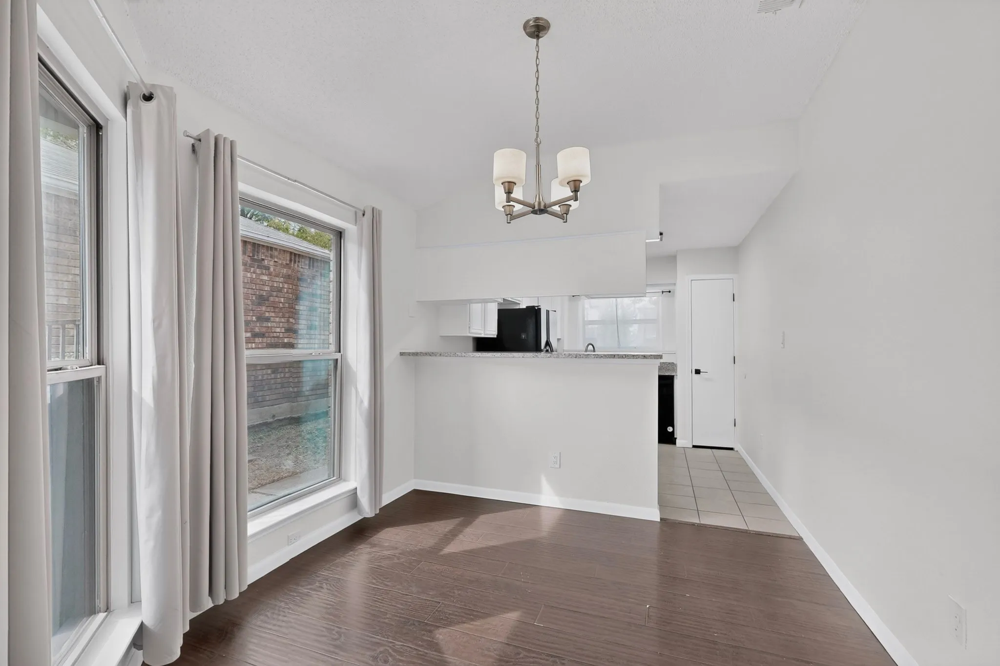 Wood-finish flooring, two large windows, overhead chandelier, white paint throughout, and a breakfast bar with a solid surface countertop