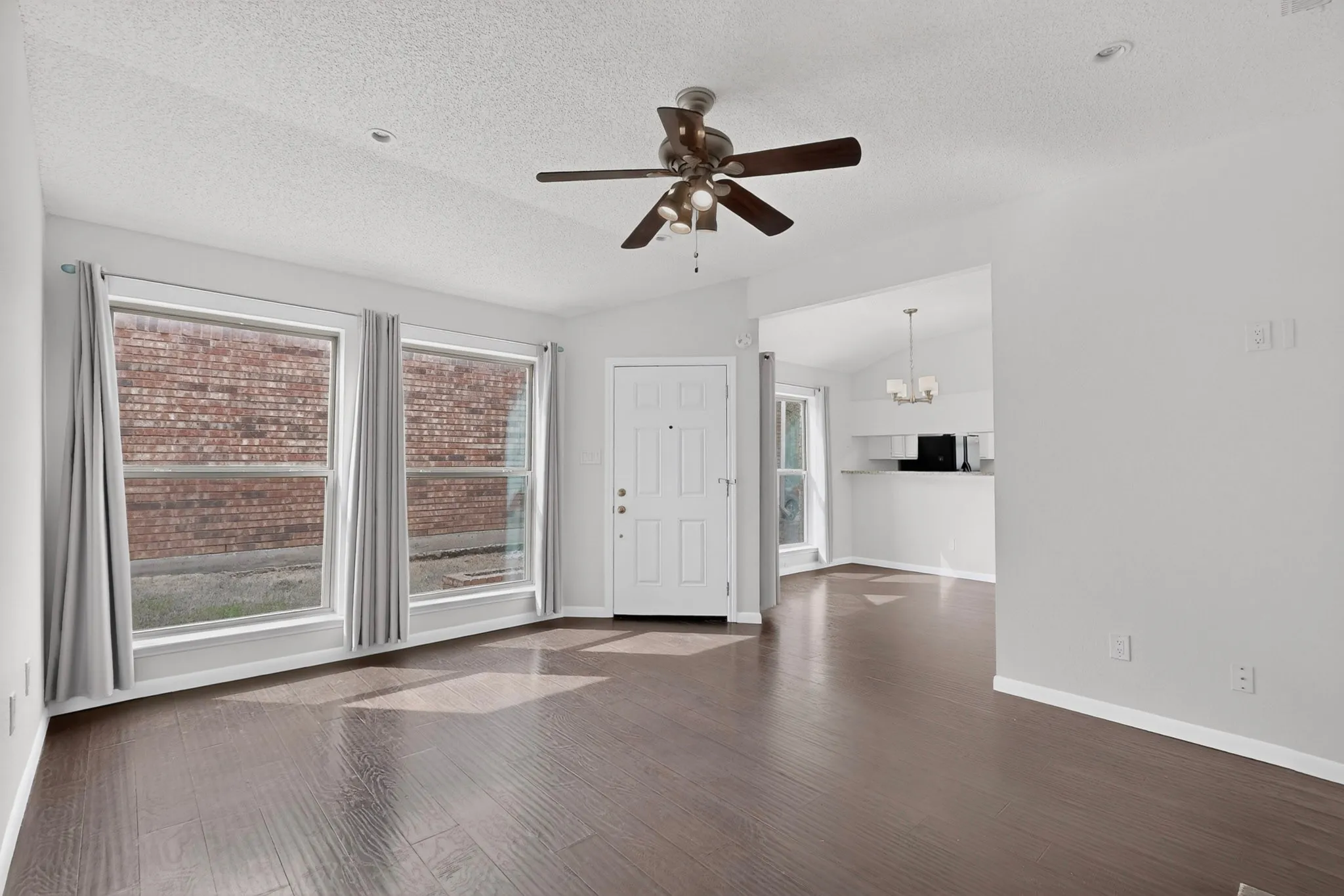 Dark wood-finish flooring, two large windows, white paneled entry door, ceiling fan with light fixture, and an open layout to an adjacent area with a chandelier