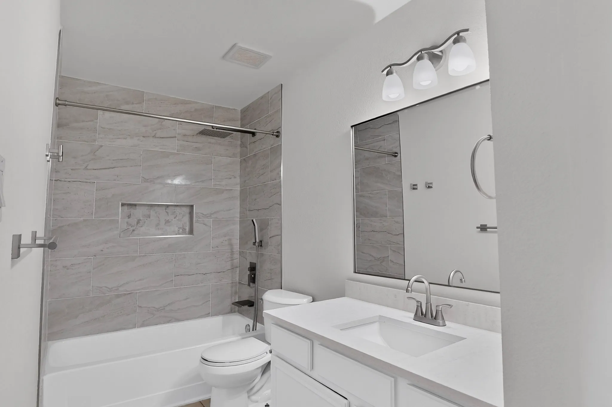 Modern bathroom featuring a vanity with a rectangular sink, a large mirror, a three-light fixture, a toilet, and a tiled shower-over-tub combination with a recessed niche