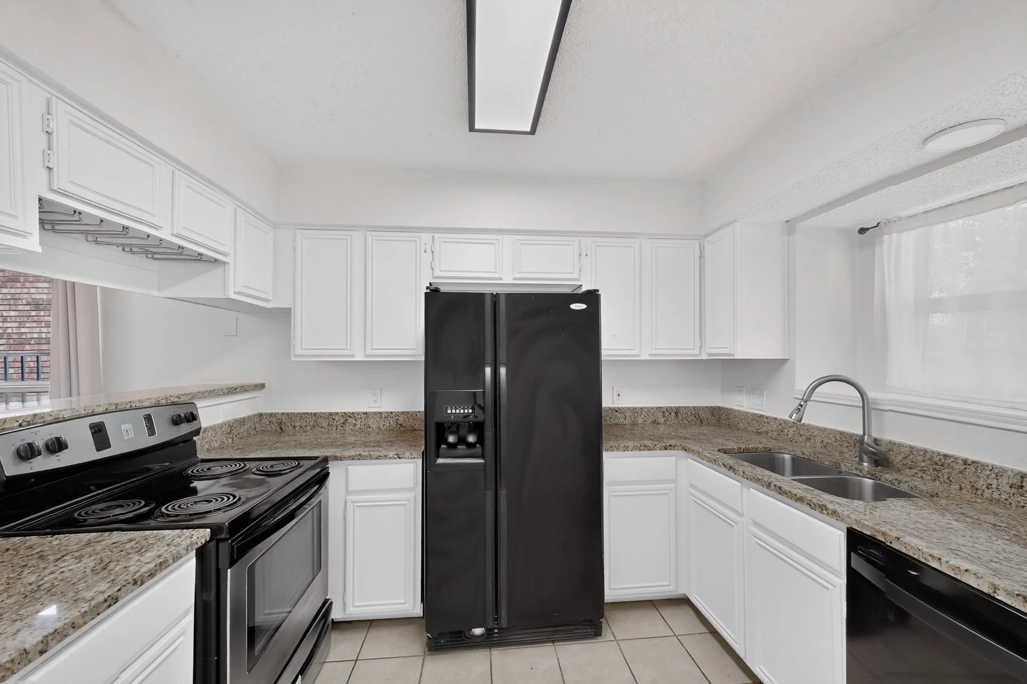 Galley kitchen featuring white cabinetry, granite countertops, stainless steel appliances including a side-by-side refrigerator and gas range, ceramic tile flooring, and a chrome pull-down faucet with dual​​‌​​​​‌​​‌‌​‌‌​​​‌‌​‌​‌​‌​​​‌​​ sinks