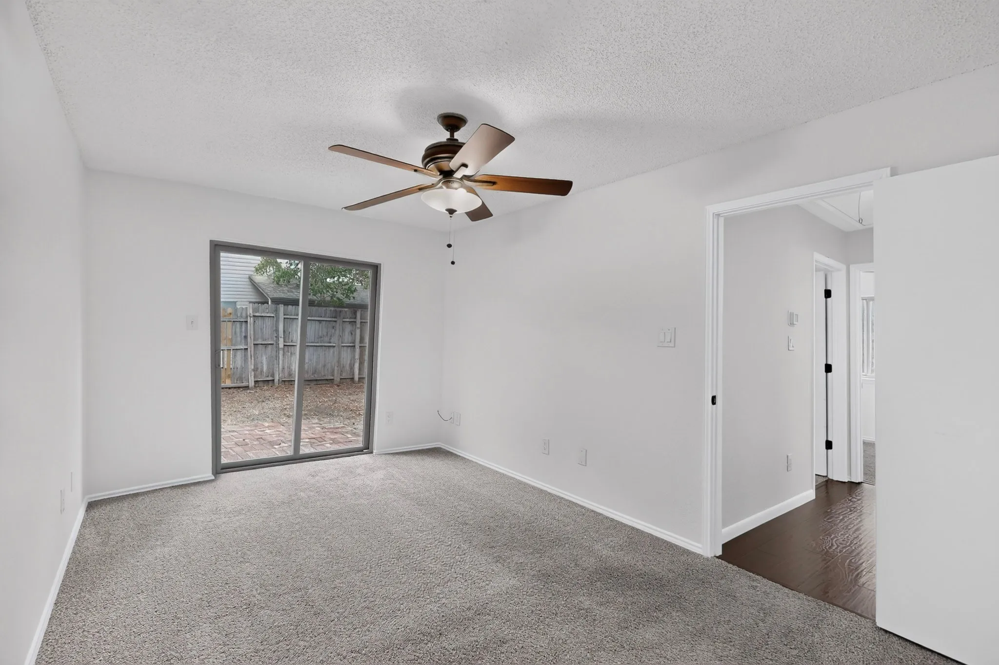 Neutral wall paint, ceiling fan with integrated lighting, sliding glass door, private outdoor area with fencing, and dark wood-finish flooring in an adjacent space