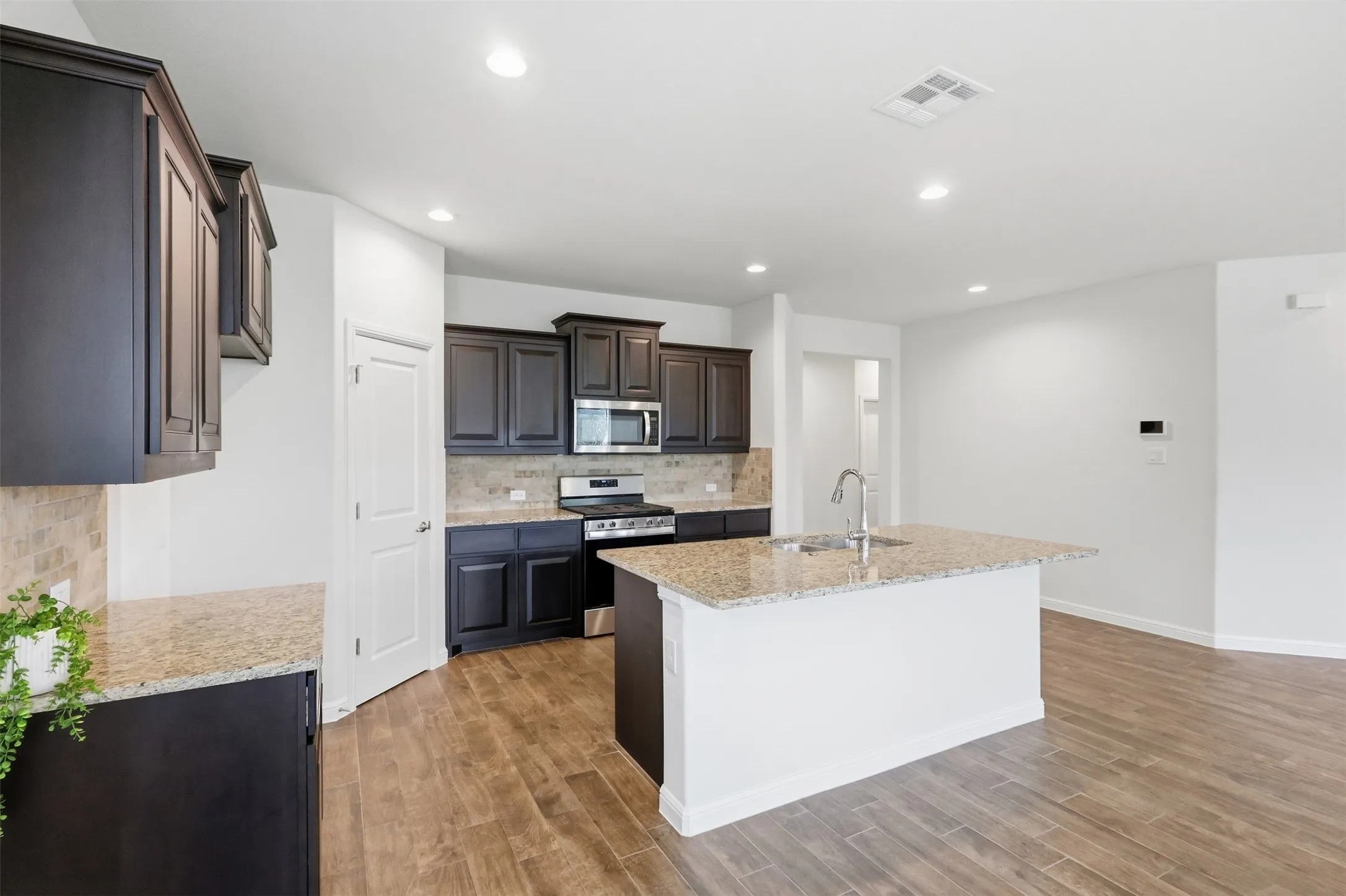 Kitchen featuring dark wood cabinetry, light-toned tiled backsplash, stainless steel appliances, and a central island with a sink and light-toned countertop