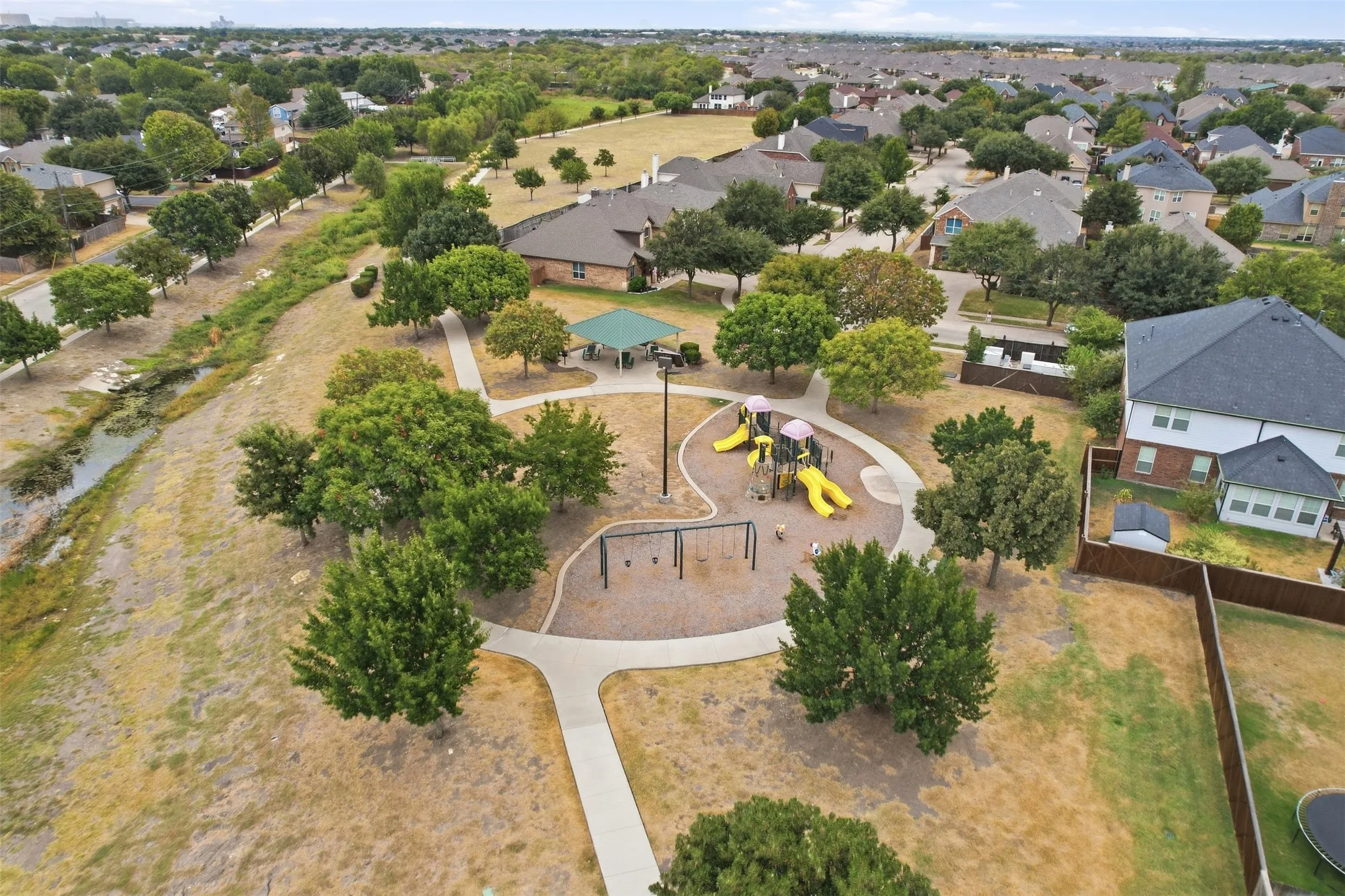 Community park with recreational equipment, mature trees, paved pathways, and a covered pavilion