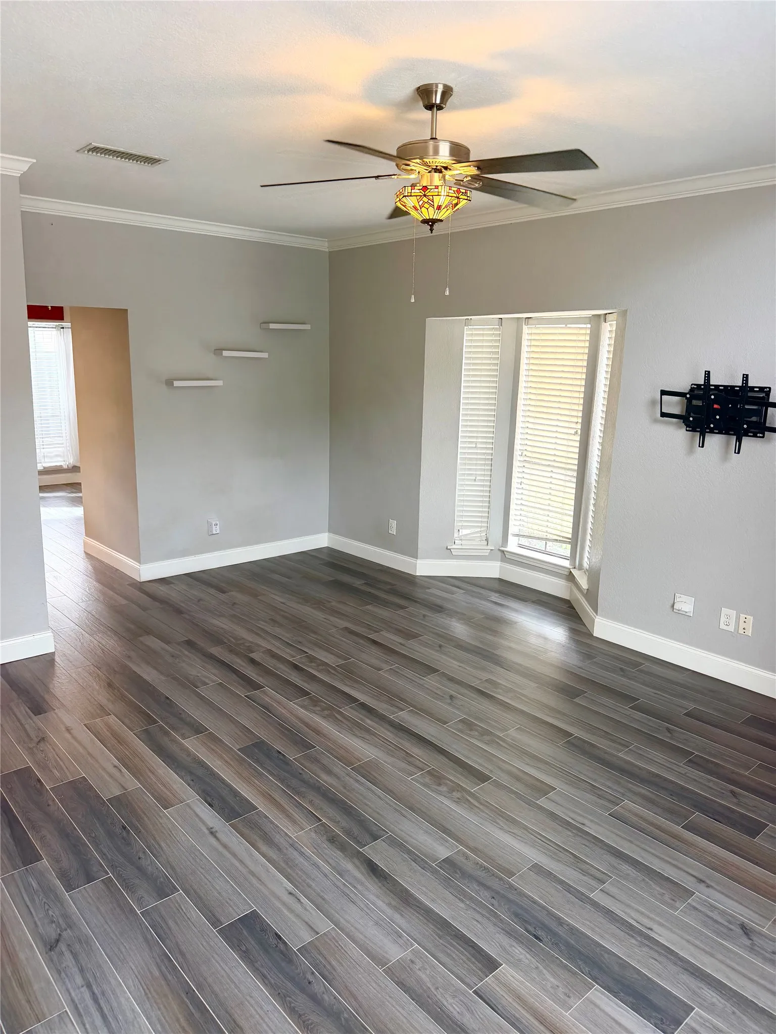 Wood-look plank flooring throughout, ceiling fan with integrated lighting, bay window with louvered blinds, and crown molding