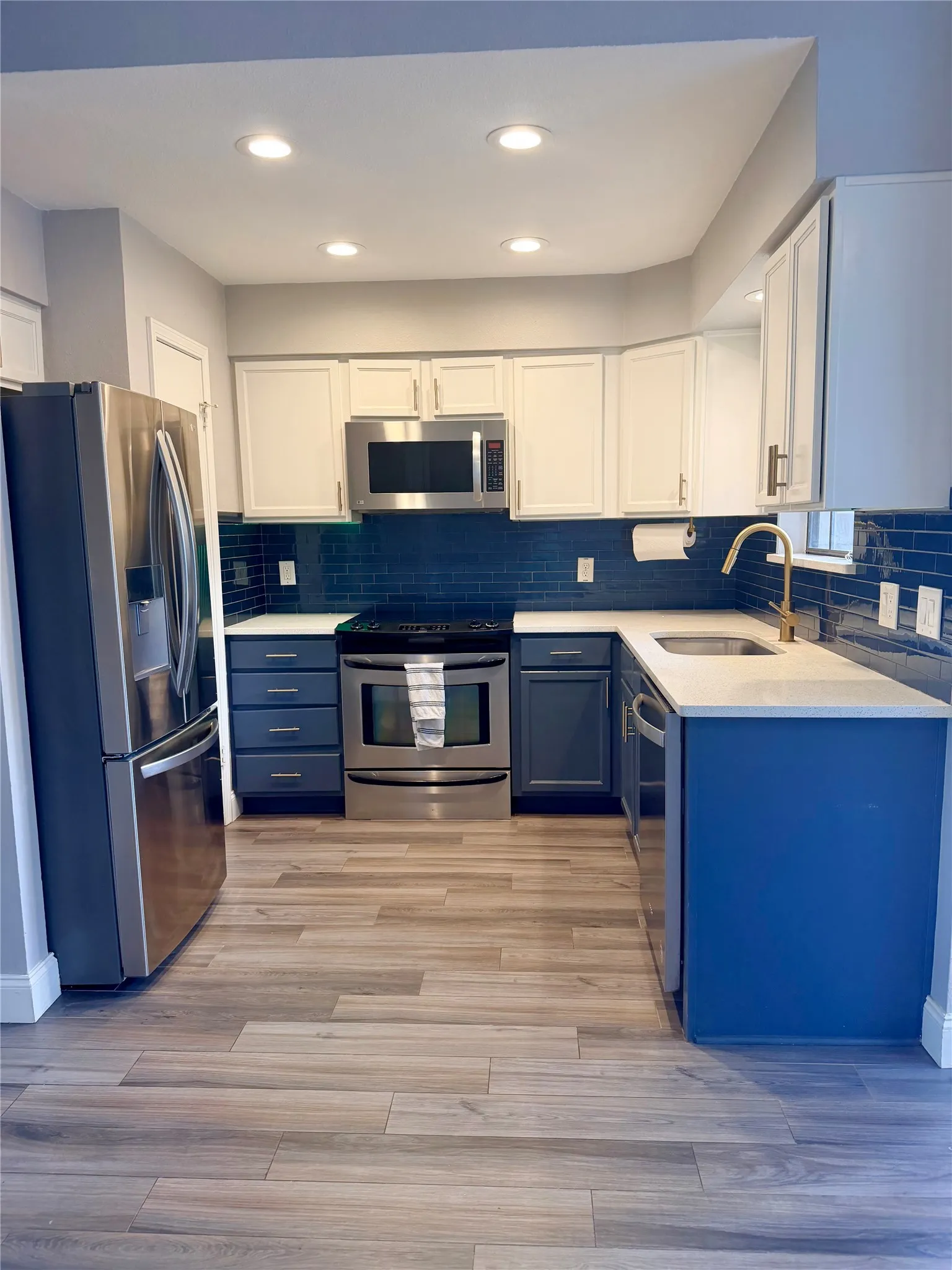 Stainless steel appliances, white upper cabinetry, blue lower cabinetry, blue subway tile backsplash, and light-toned wood-finish flooring