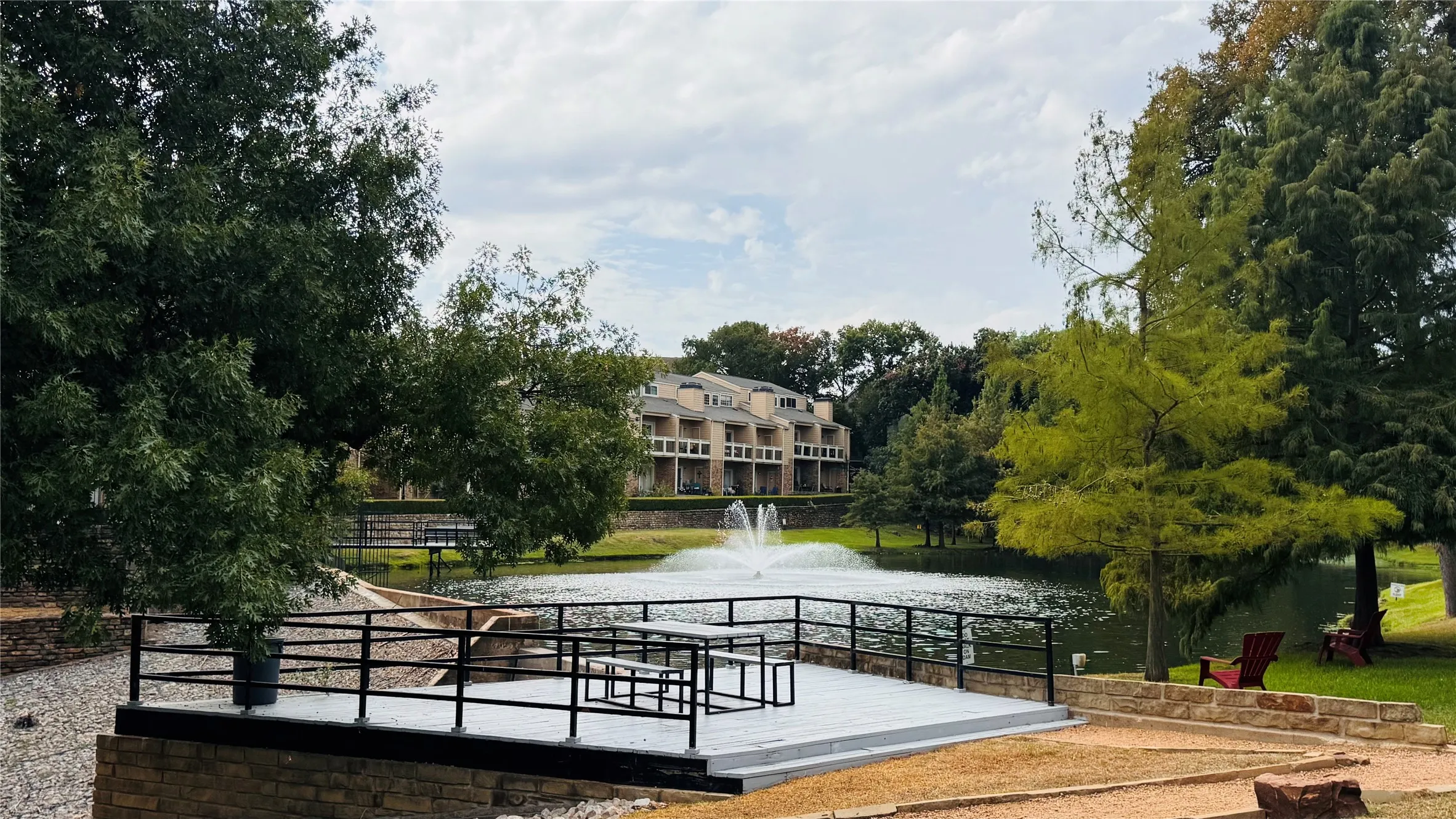 Deck with black railings and a built-in bench, offering a serene water feature with a fountain, surrounded by mature trees and lush green landscaping