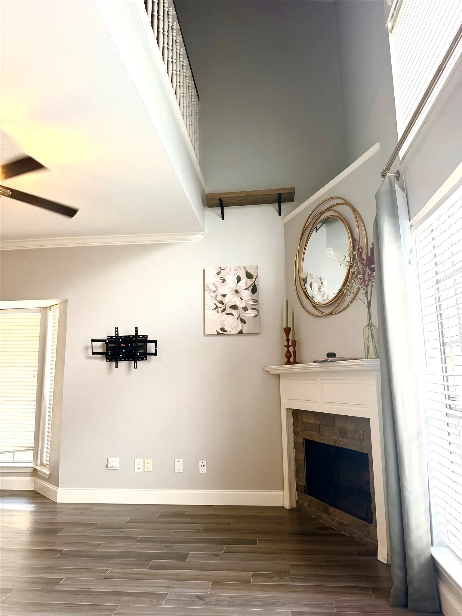 Wood-finish flooring, fireplace with stone surround and white mantel, crown molding, double-height ceiling, and built-in shelving