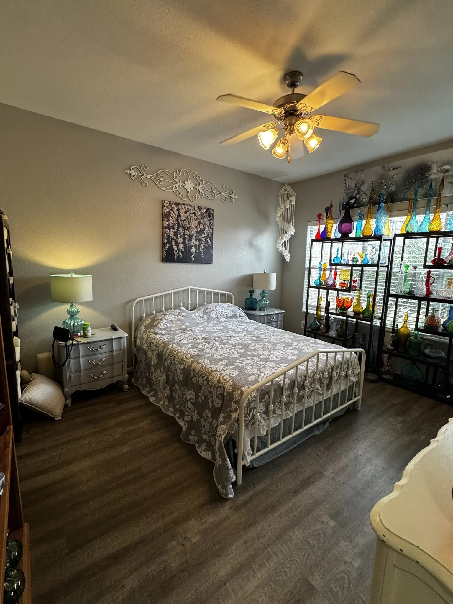 Comfortable bedroom retreat showcasing a metal bed frame, wood-finish flooring, ceiling fan with integrated lighting, bedside nightstand with task lighting, and built-in shelving with display storage along the adjacent wall