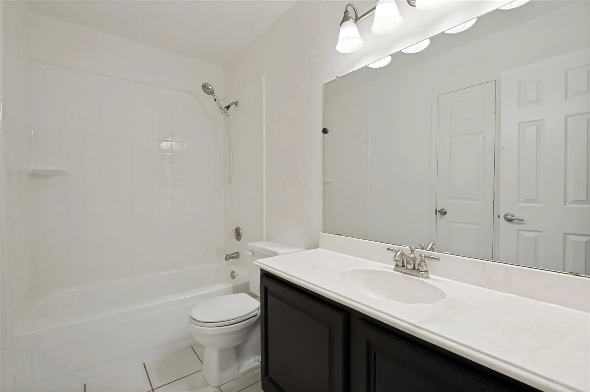 White tiled flooring, dark wood-finish vanity with a white countertop, recessed sink, and a framed mirror with overhead lighting