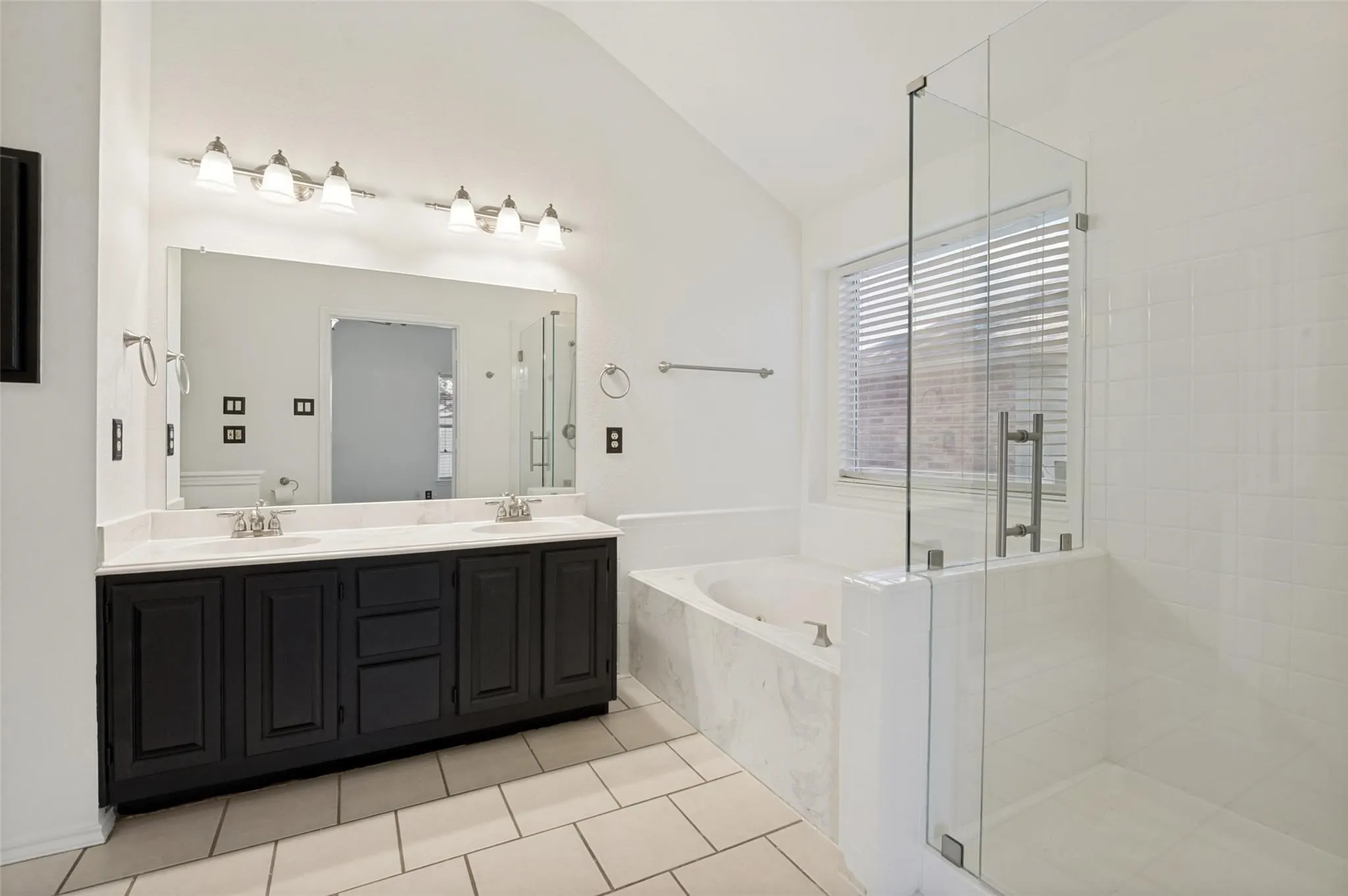 Dual vanity with dark wood-finish cabinetry and light-toned countertop, large frameless mirror, glass-enclosed shower with white tiled walls, and a built-in soaking tub