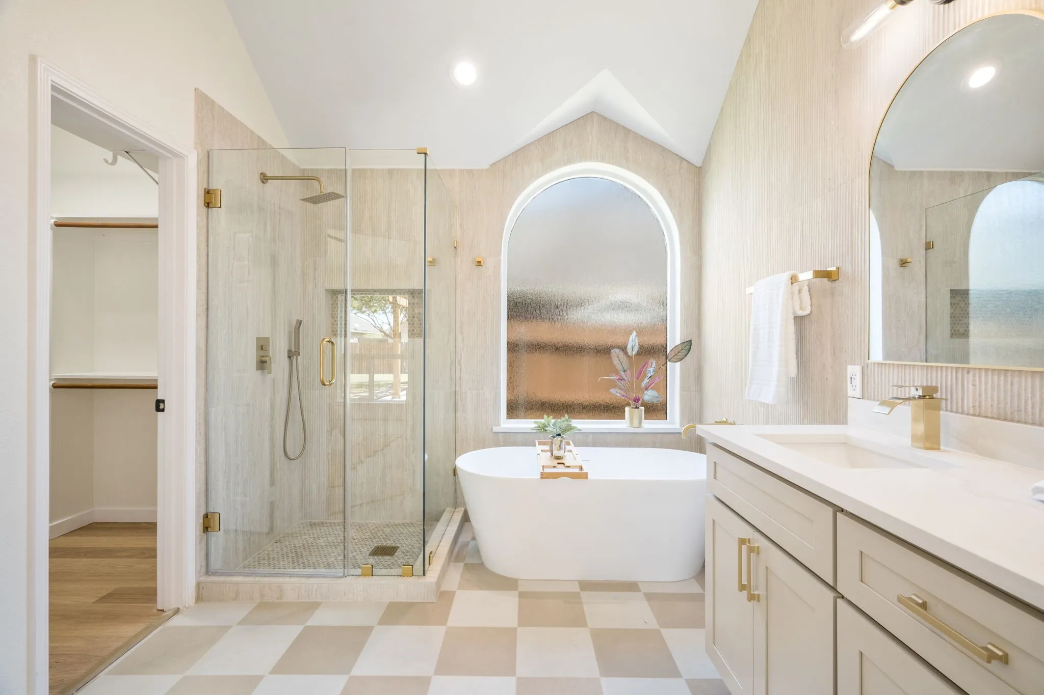 Freestanding soaking tub with arched window, glass-enclosed shower, dual vanity with undermount sinks, and checkered tile flooring