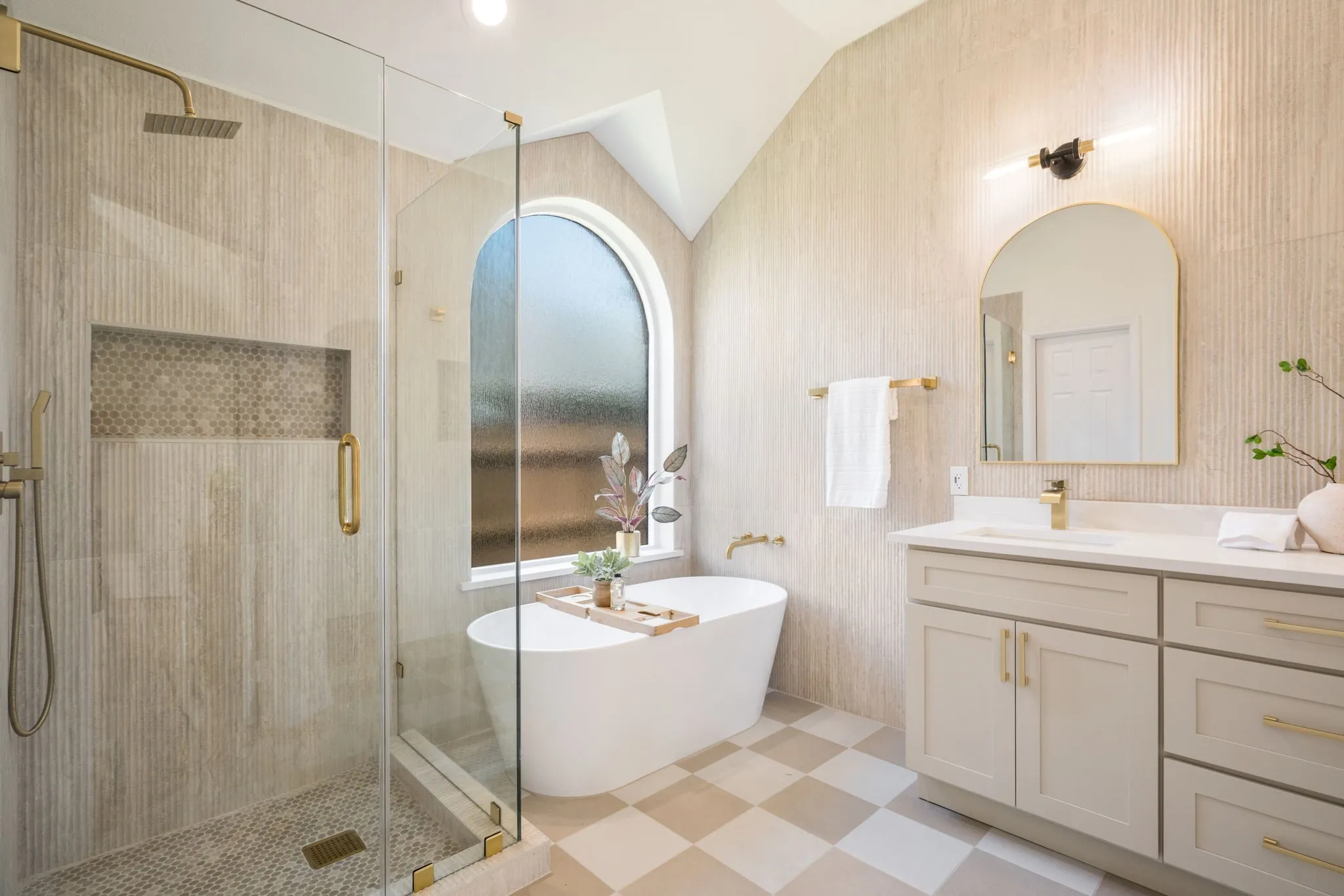 Glass-enclosed shower with a recessed niche, free-standing soaking tub, arched window, single vanity with a white countertop, and checkered floor tiling