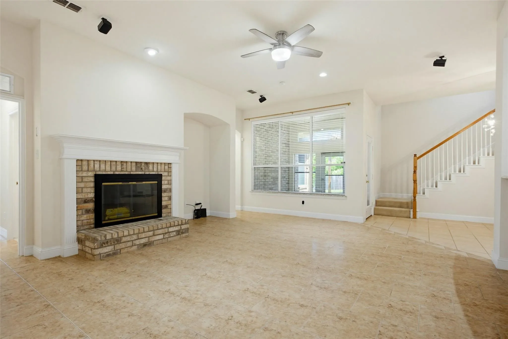 Neutral-toned tile flooring, brick-faced fireplace with white mantel, ceiling fan with integrated lighting, recessed lighting, and a staircase with white spindles and wood handrail