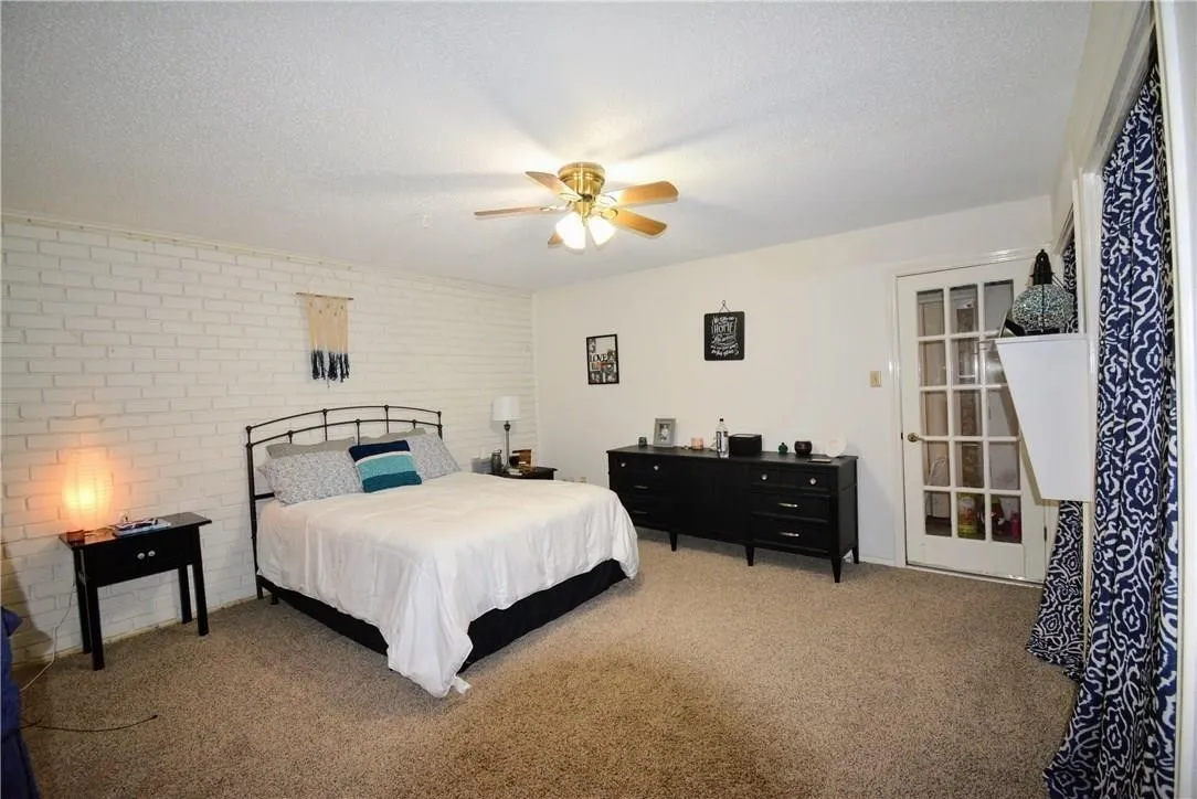 Carpeted room featuring a white painted brick accent wall, ceiling fan with light fixture, and a glass-paned door