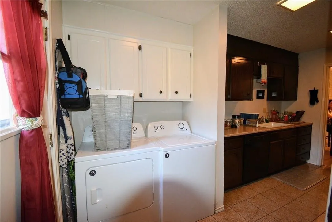 Laundry area with stacked appliances, upper white cabinetry, and adjacent kitchen with dark wood-finish cabinetry, light-toned countertop, and tiled flooring