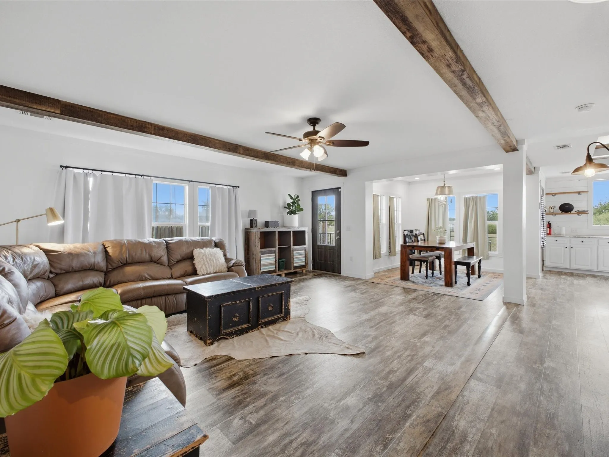 Spacious open-concept living area featuring exposed wood beams, wood-finish flooring, ample natural light, and a ceiling fan