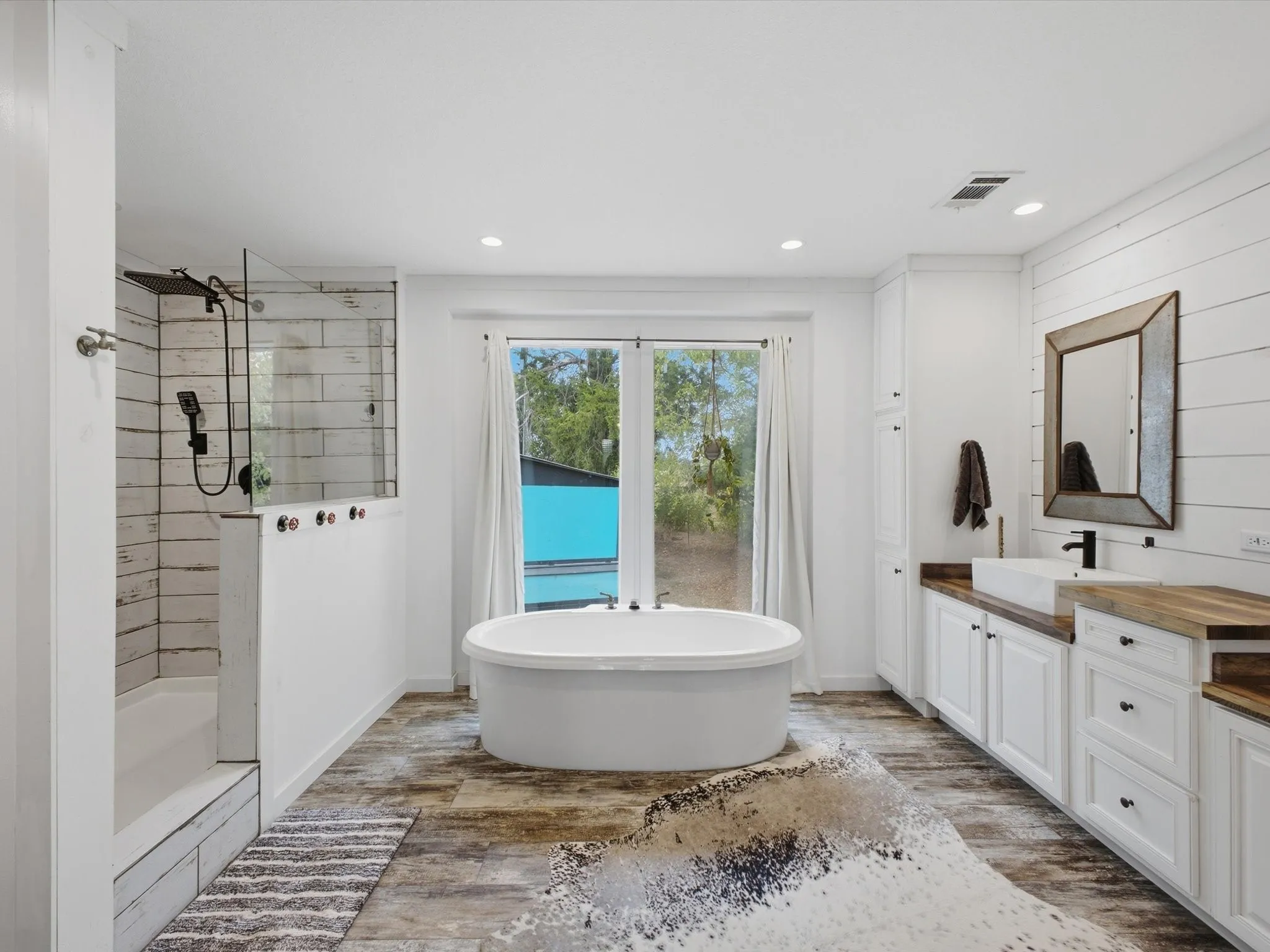 Spa-inspired primary bathroom featuring a freestanding soaking tub, walk-in shower with subway tile surround and rainfall showerhead, dual vanity with wood-finish countertop, white cabinetry with hardware accents, and large-format tile flooring
