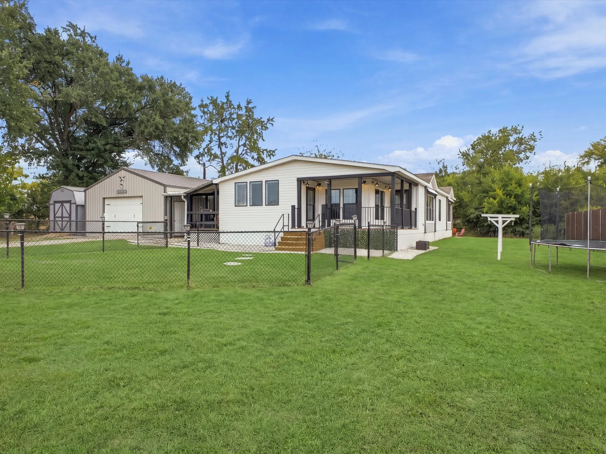 Manicured lawn, full chain-link fencing, detached garage with roll-up door, covered patio with railing, and a secondary storage shed