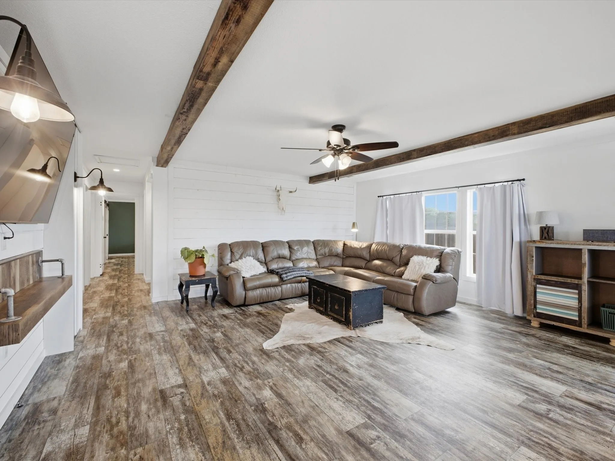 Spacious living area featuring wood-finish flooring, exposed ceiling beams, a ceiling fan, and white shiplap accent wall