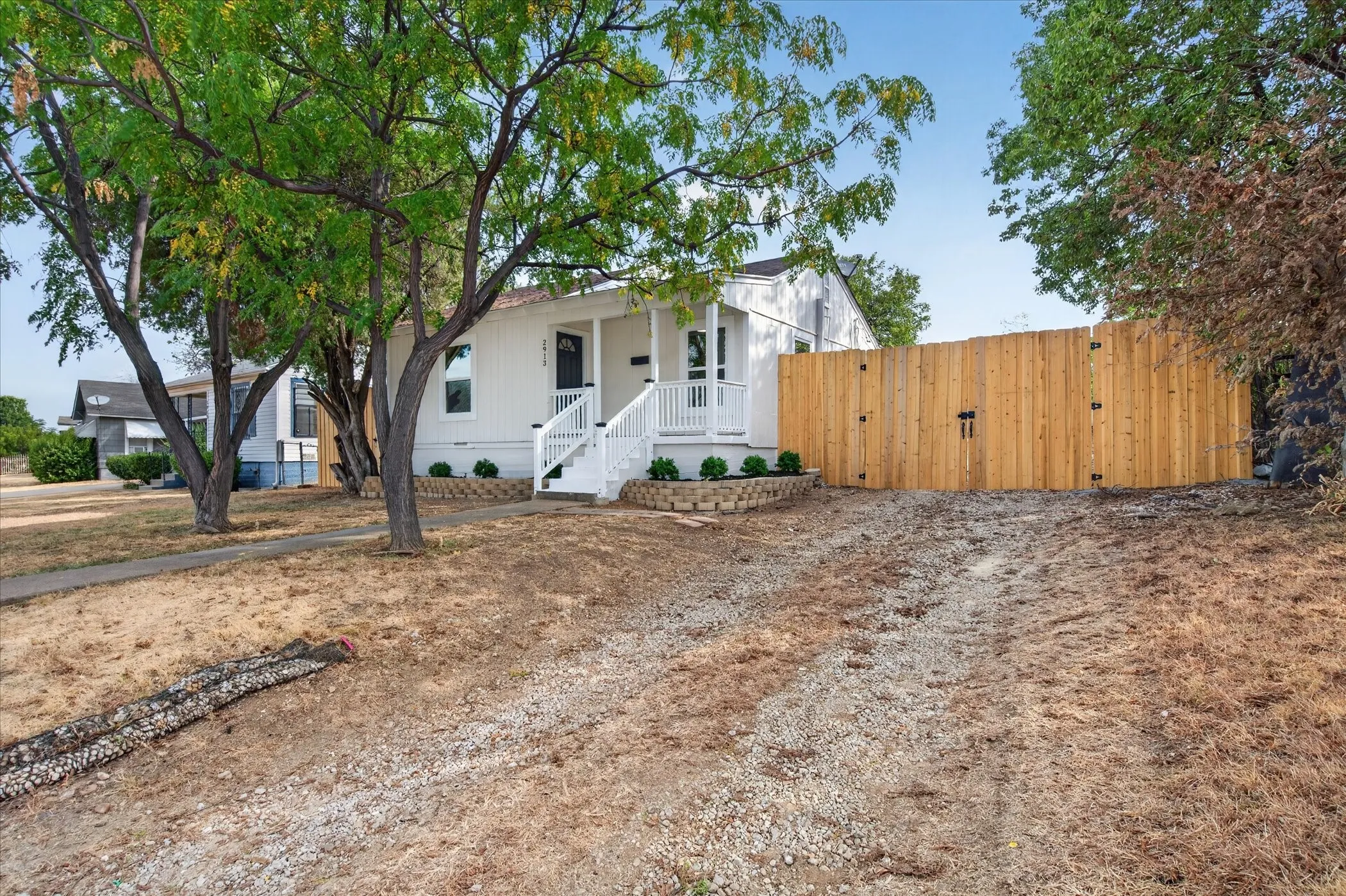 White exterior with dark trim, front porch with white railing and steps, mature trees, and a wooden privacy fence with gate