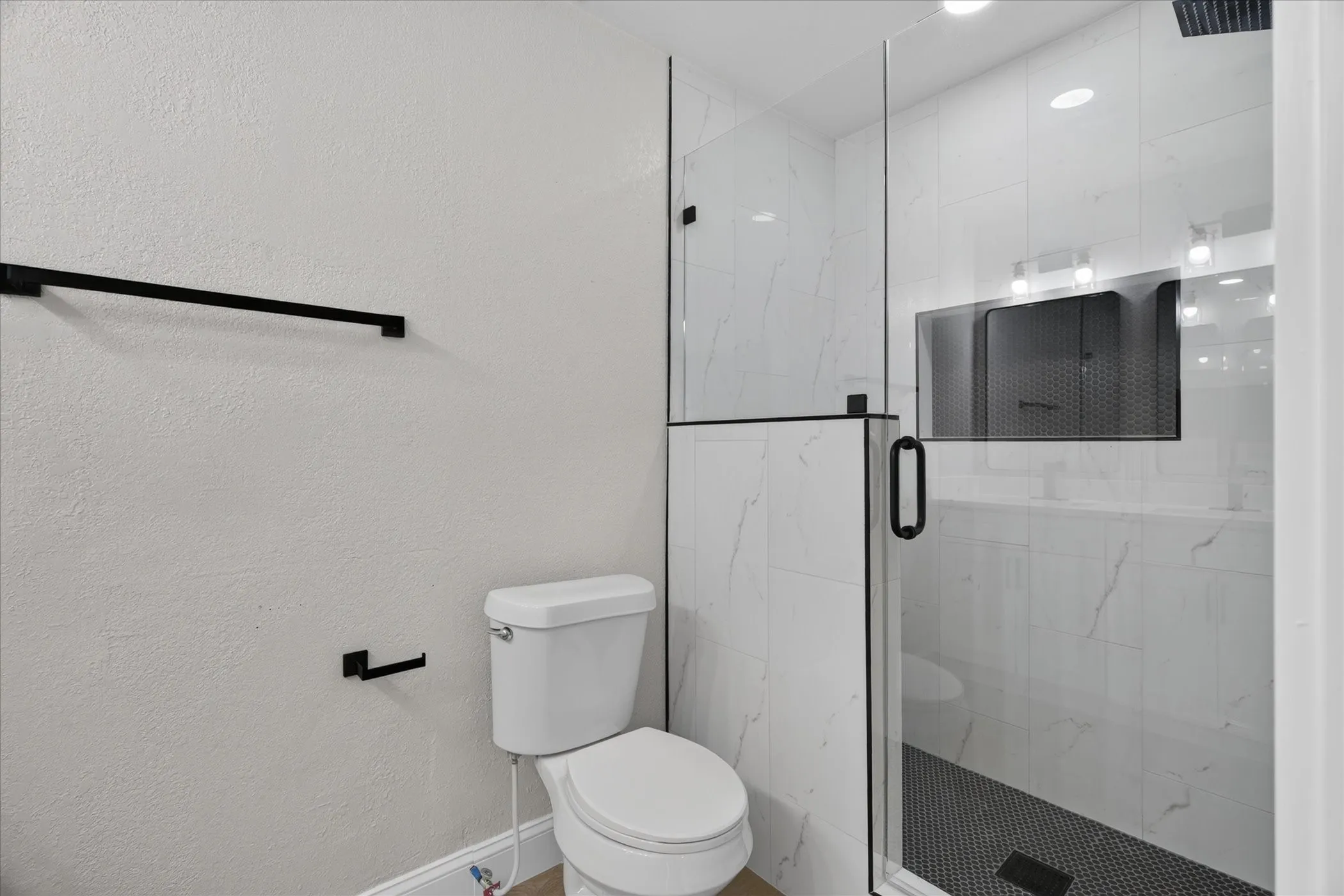 Frameless glass shower enclosure with matte black hardware, white tile with subtle veining, recessed shower niche, and a black penny tile shower floor