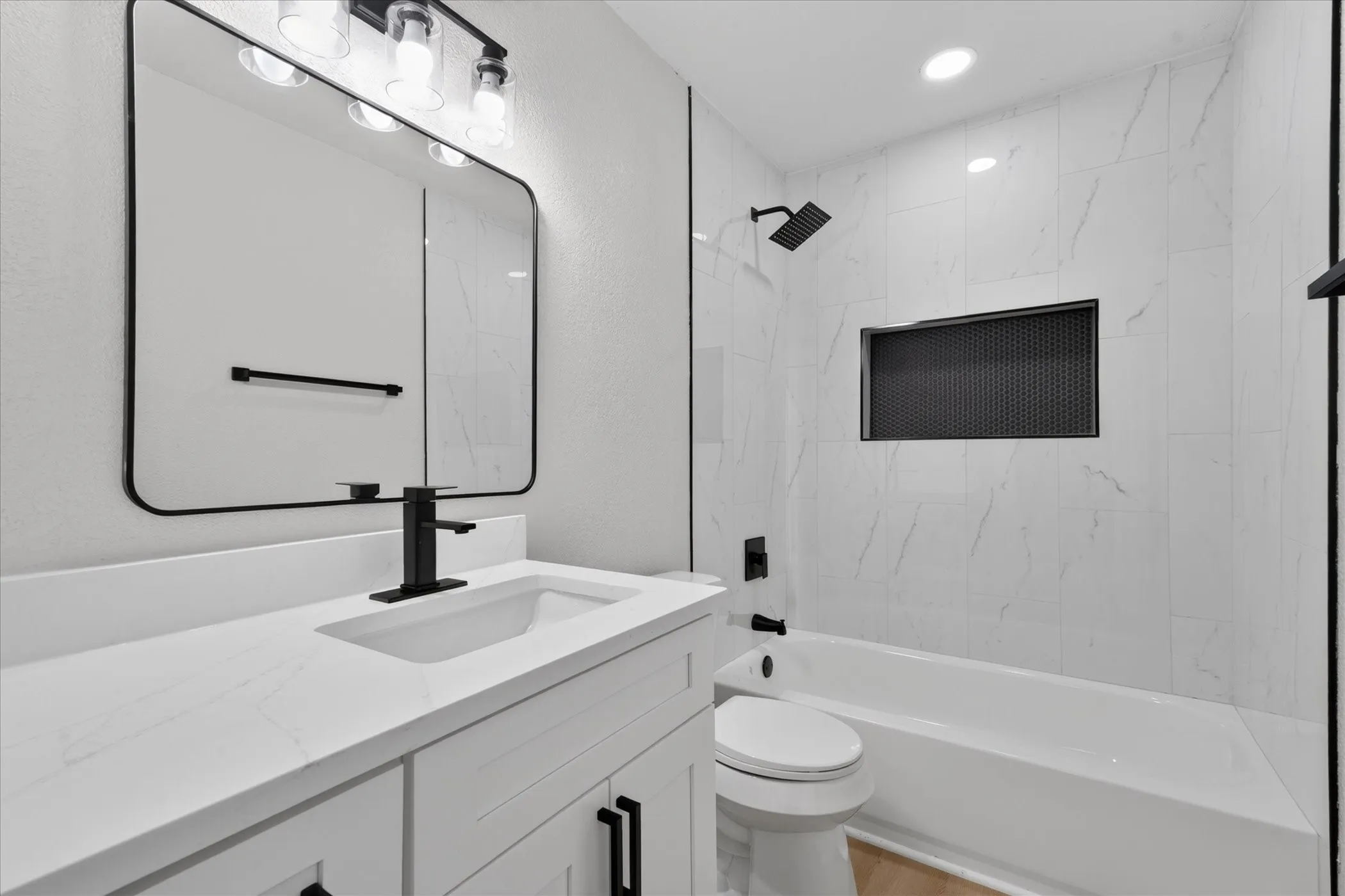Bathroom featuring a white vanity with integrated sink, matte black fixtures, white tiled tub surround with recessed niche, and a contemporary mirror