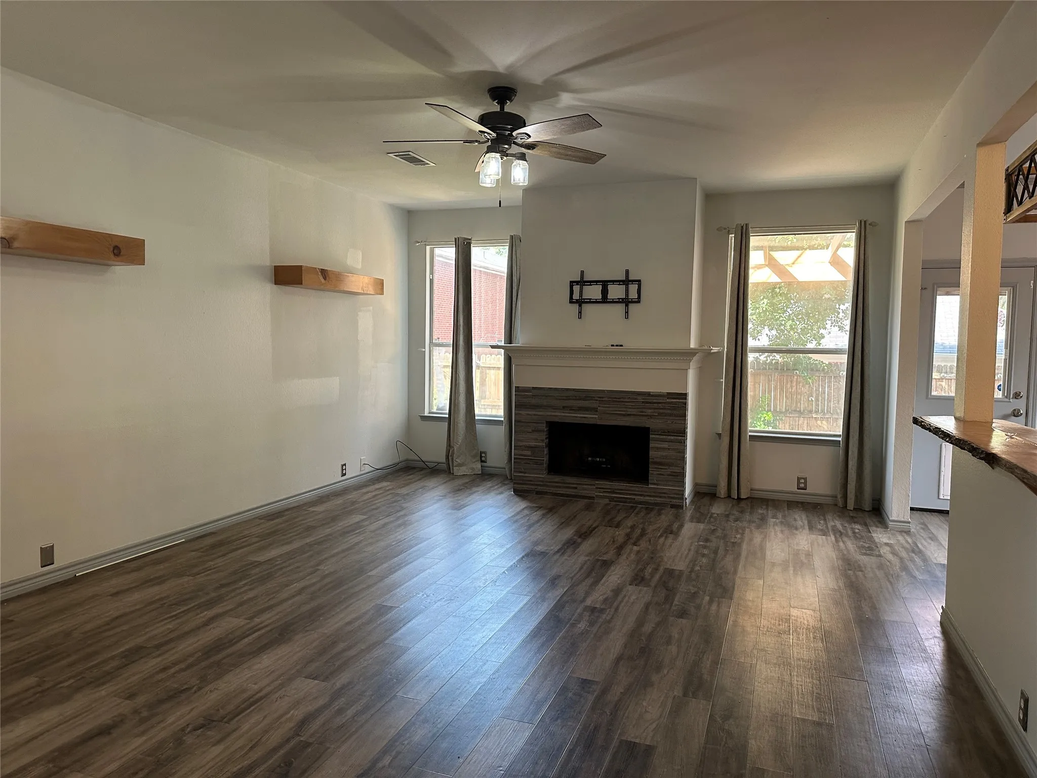 Spacious living area featuring wood-finish flooring, a ceiling fan with integrated lighting, a fireplace with a stone surround, and built-in floating shelves
