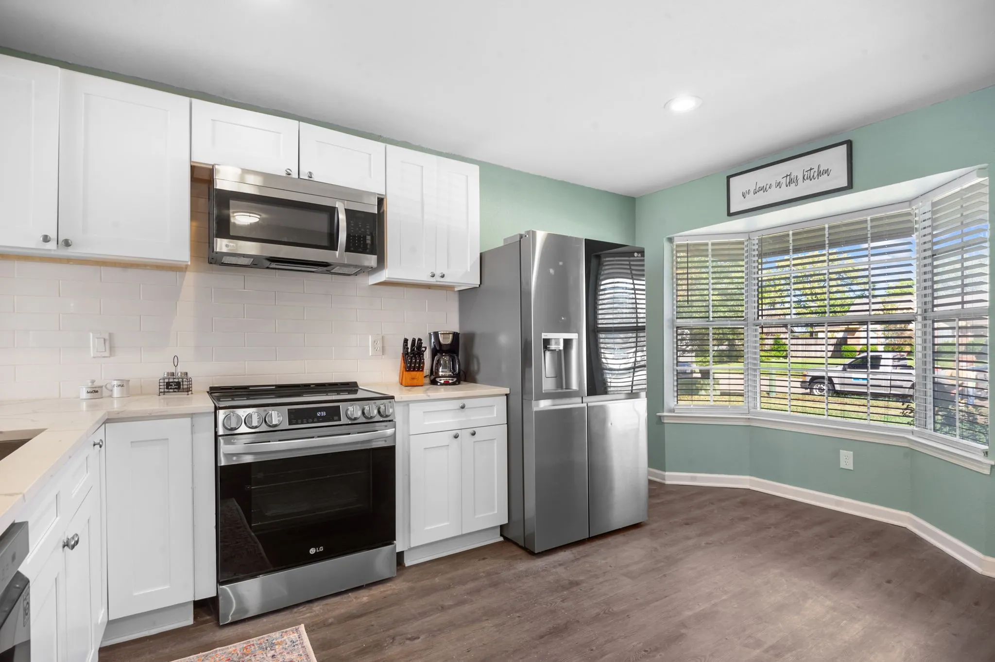 Modern kitchen featuring white cabinetry, stainless steel appliances, a subway tile backsplash, wood-finish flooring, and a bay window