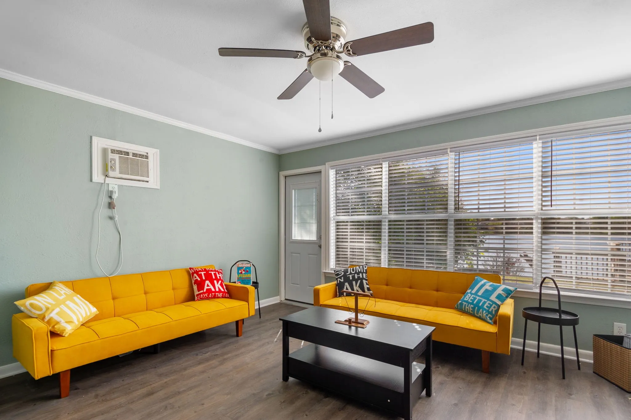 Wood-finish flooring, ceiling fan, multiple windows with blinds, and a white entry door with glass paneling