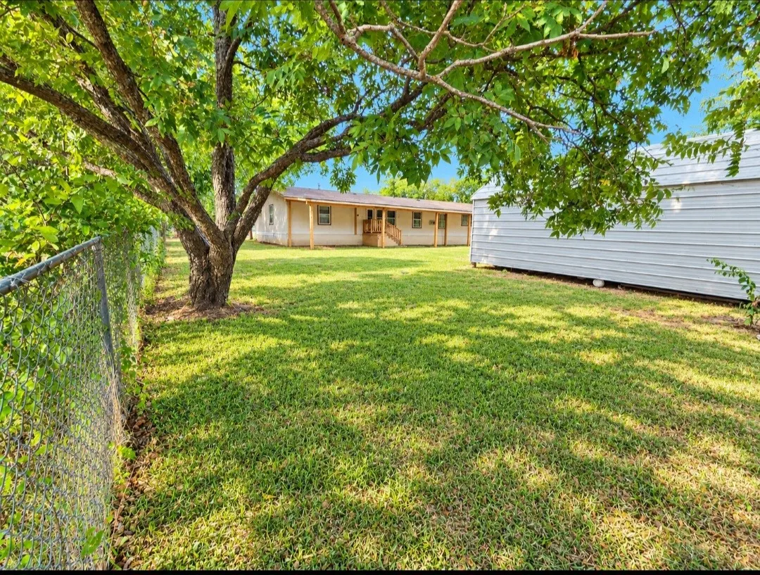 Expansive grassy yard with mature trees, a chain-link fence, and a storage shed