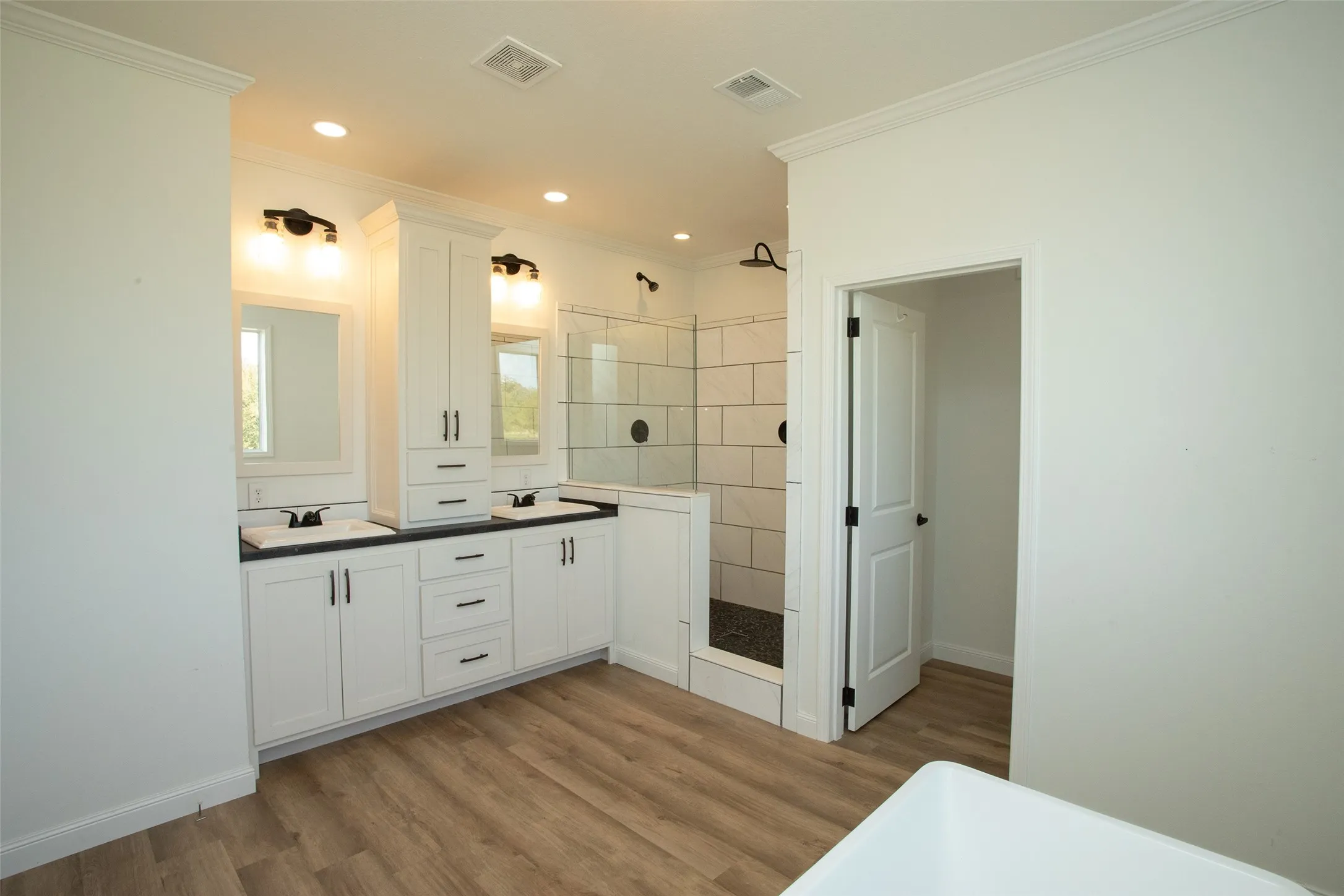 Dual vanities with integrated sinks, wood-finish flooring, a glass-enclosed shower with subway tile, white shaker-style cabinetry, and recessed lighting
