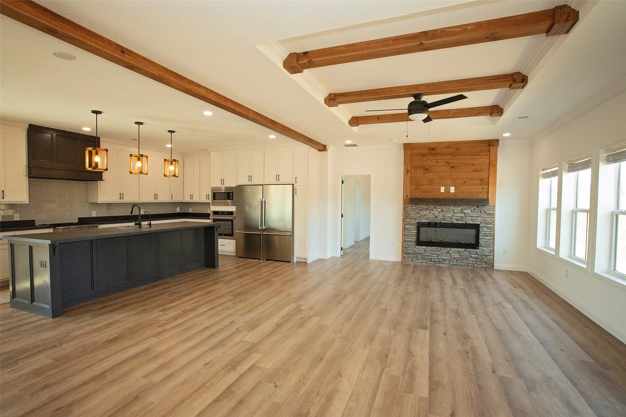 Open-concept living area with wood-finish flooring, a kitchen island, stainless steel appliances, a stone-face fireplace, and a tray ceiling with wood beams