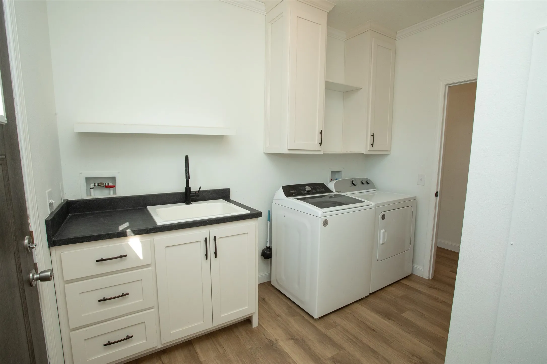Laundry area featuring wood-finish flooring, white cabinetry, a utility sink with a black faucet, and a dark countertop
