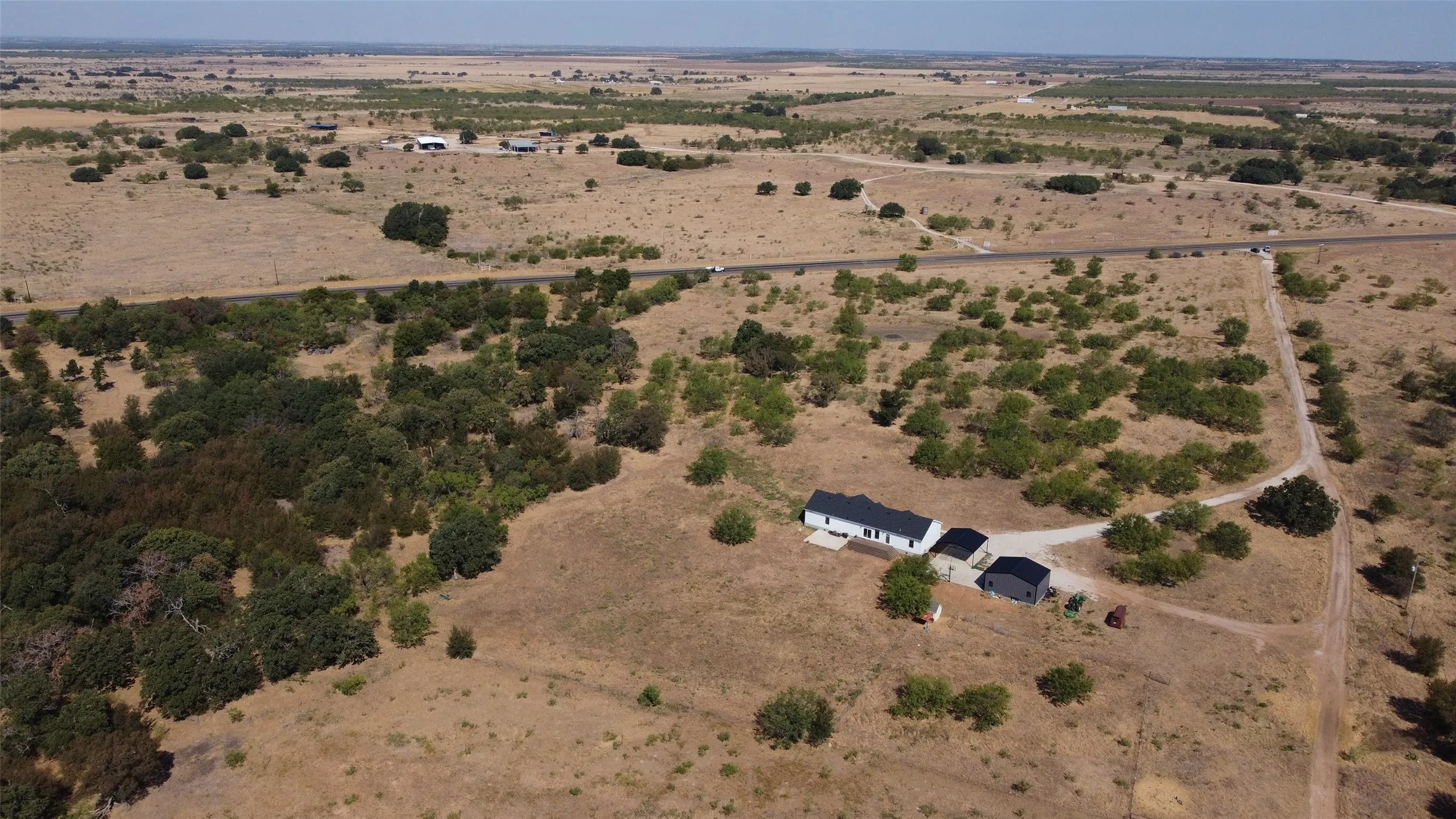 Expansive rural property featuring a white residence, a dark-colored accessory structure, and a broad unpaved driveway