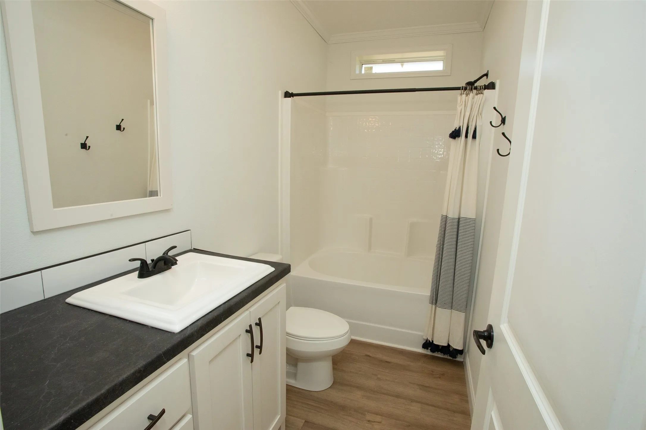 White vanity with a dark countertop and white sink, wood-finish flooring, white bathtub and shower surround, and a clerestory window