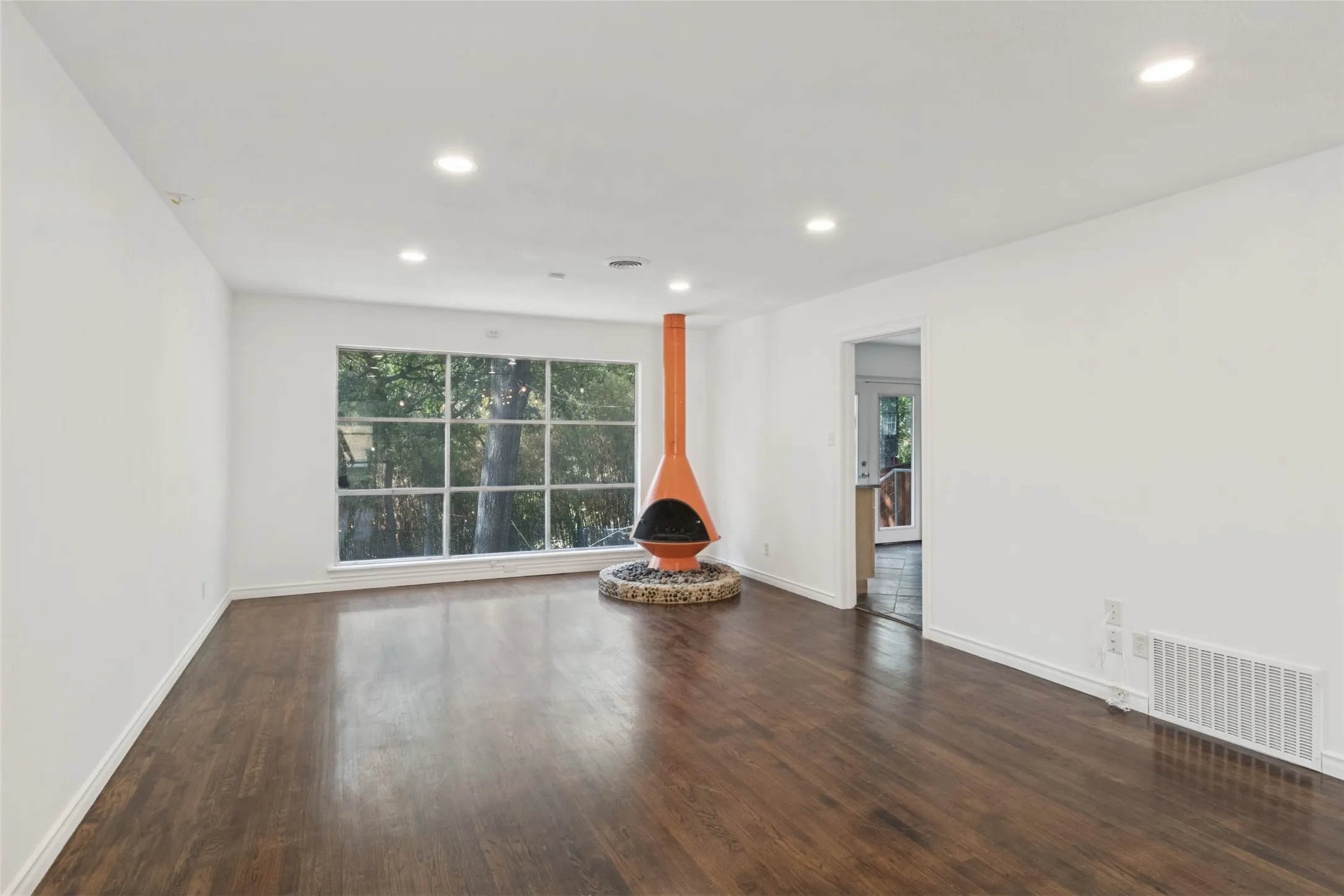 Spacious room featuring wood-finish flooring, recessed lighting, a large picture window, and a unique freestanding fireplace with a stone-clad base