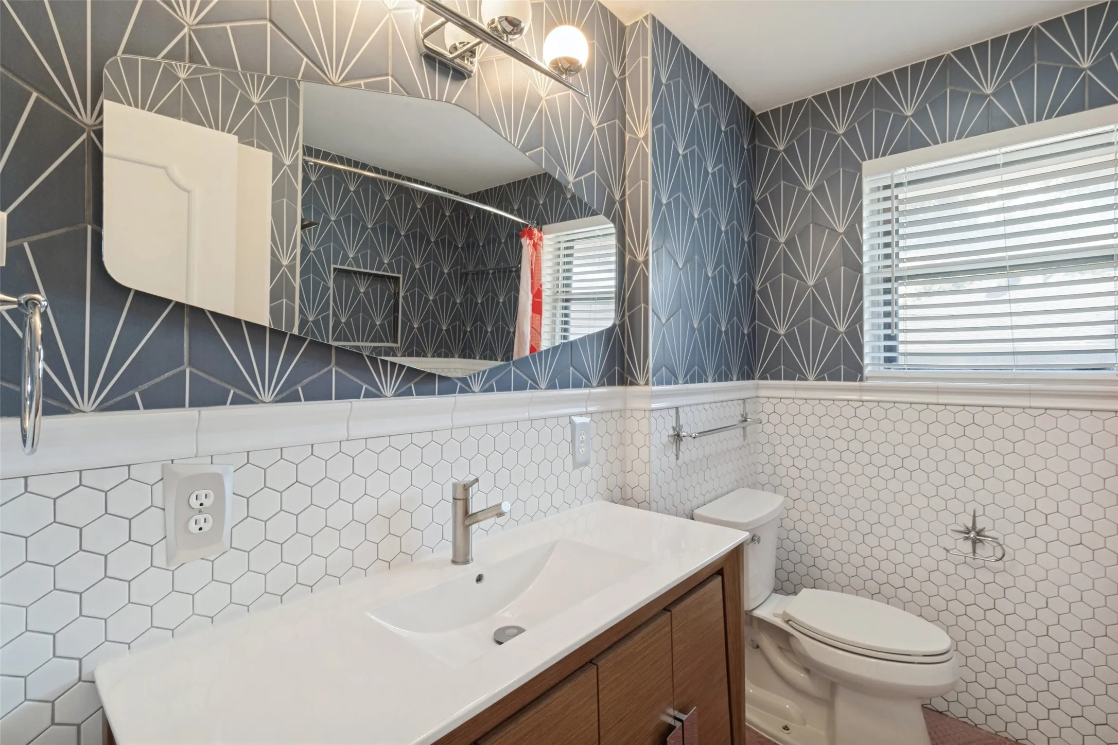 White hexagonal tile wainscoting, wood-finish vanity with integrated sink, contemporary wall-mounted light fixture, and decorative wall tiling