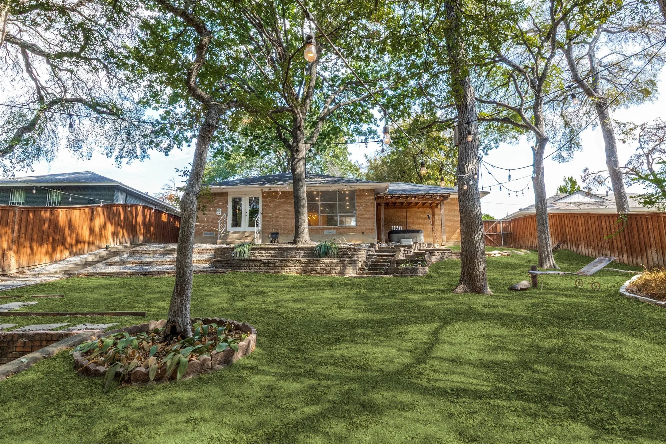 Expansive backyard with a manicured lawn, established trees, tiered stone landscaping, and a covered patio