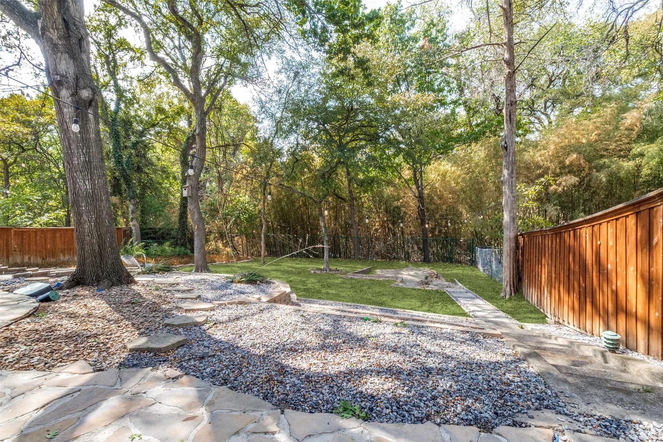 Expansive backyard featuring a stone patio, gravel landscaping, mature trees, a manicured lawn, and a privacy fence